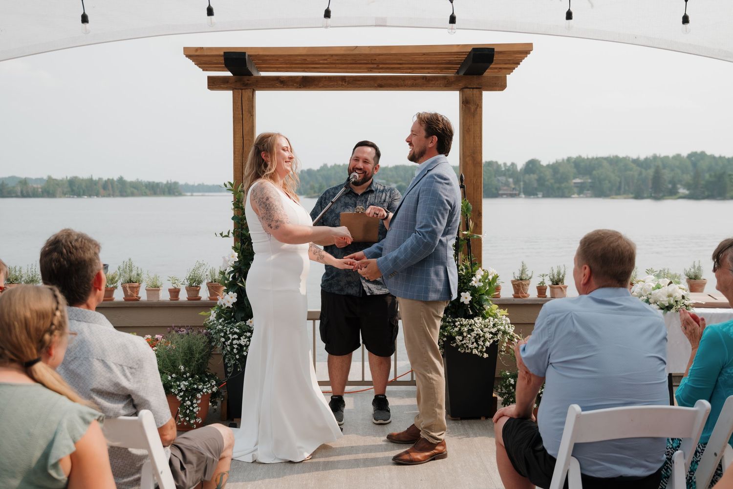 A lakeside wedding ceremony takes place under a wooden pergola with string lights and water views.