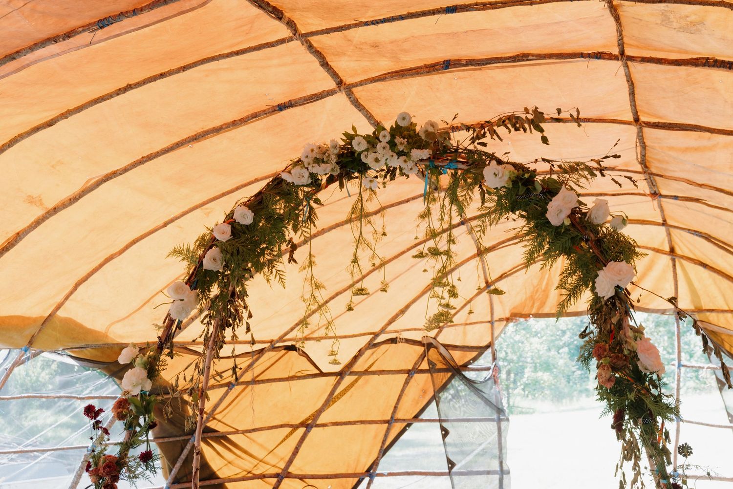 Floral arch with greenery and pink roses under a golden tent canopy at an outdoor wedding ceremony.