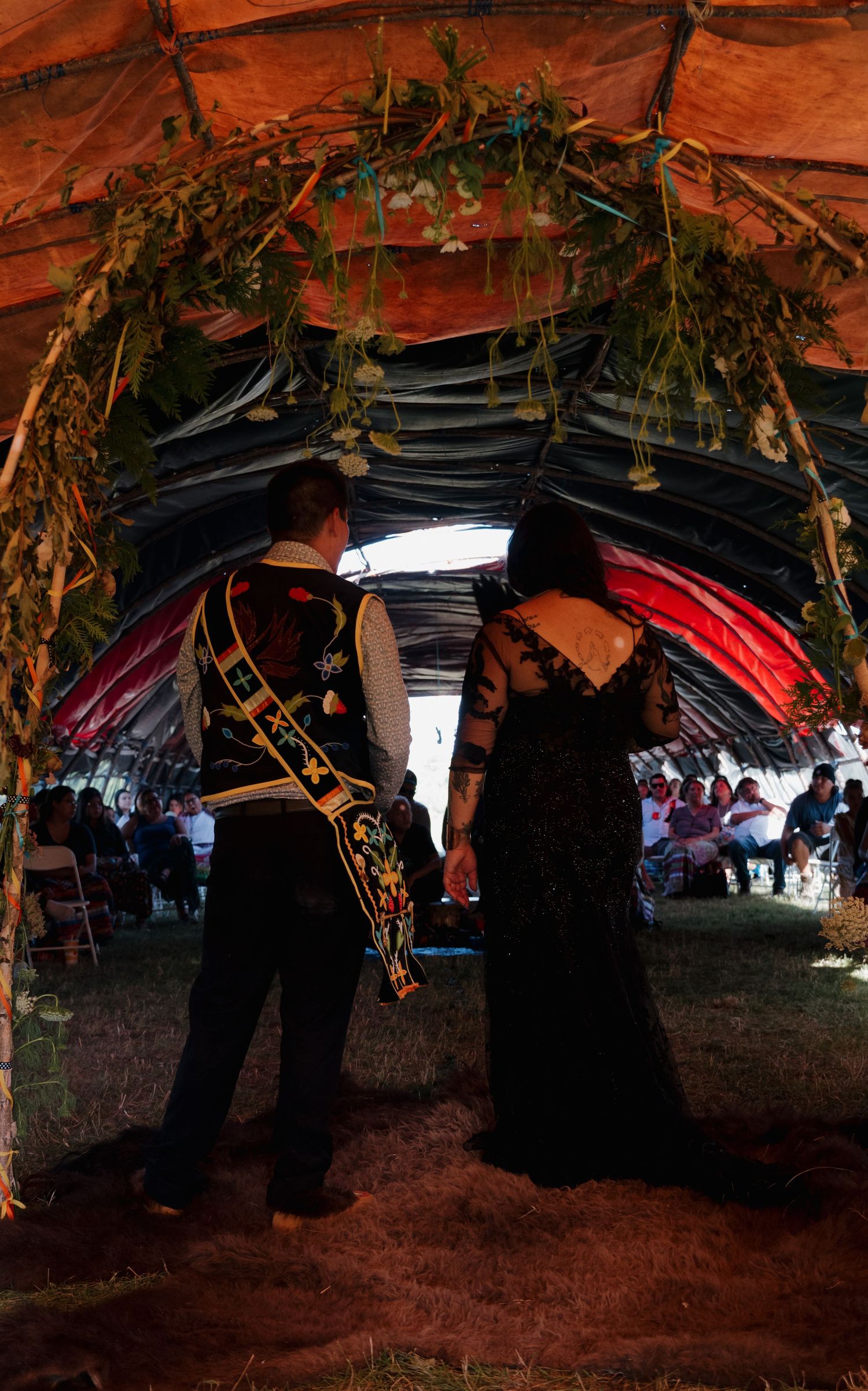 Couple in Indigenous wedding ceremony walks hand-in-hand under floral arch inside traditional dome structure with seated guests.