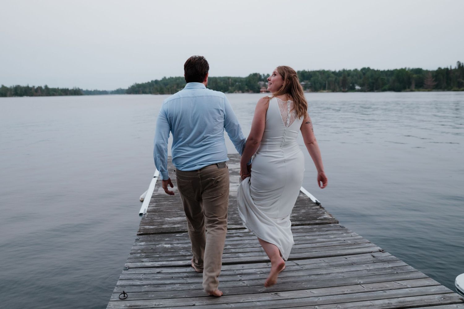 Couple walking hand in hand on a wooden pier with a lake view.