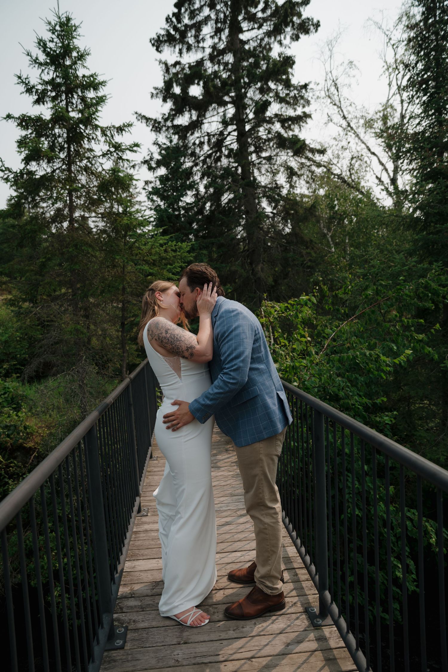 A romantic moment captured on a wooden bridge surrounded by evergreen trees in a natural outdoor setting.