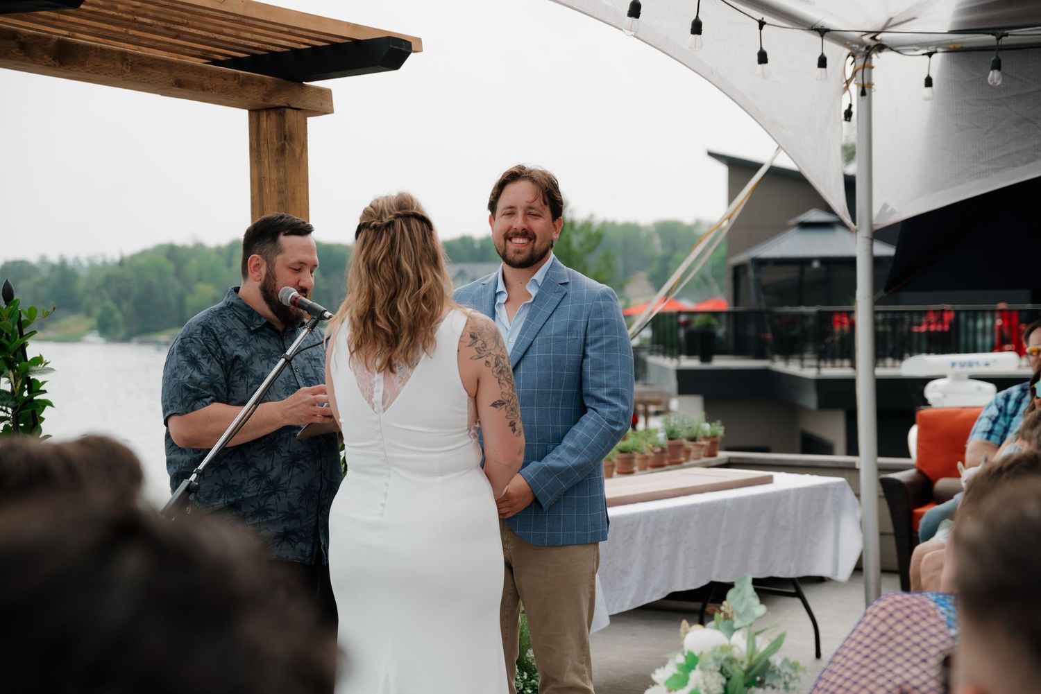 Smiling groom exchanges vows with bride during an outdoor lakeside wedding ceremony with guests seated nearby.