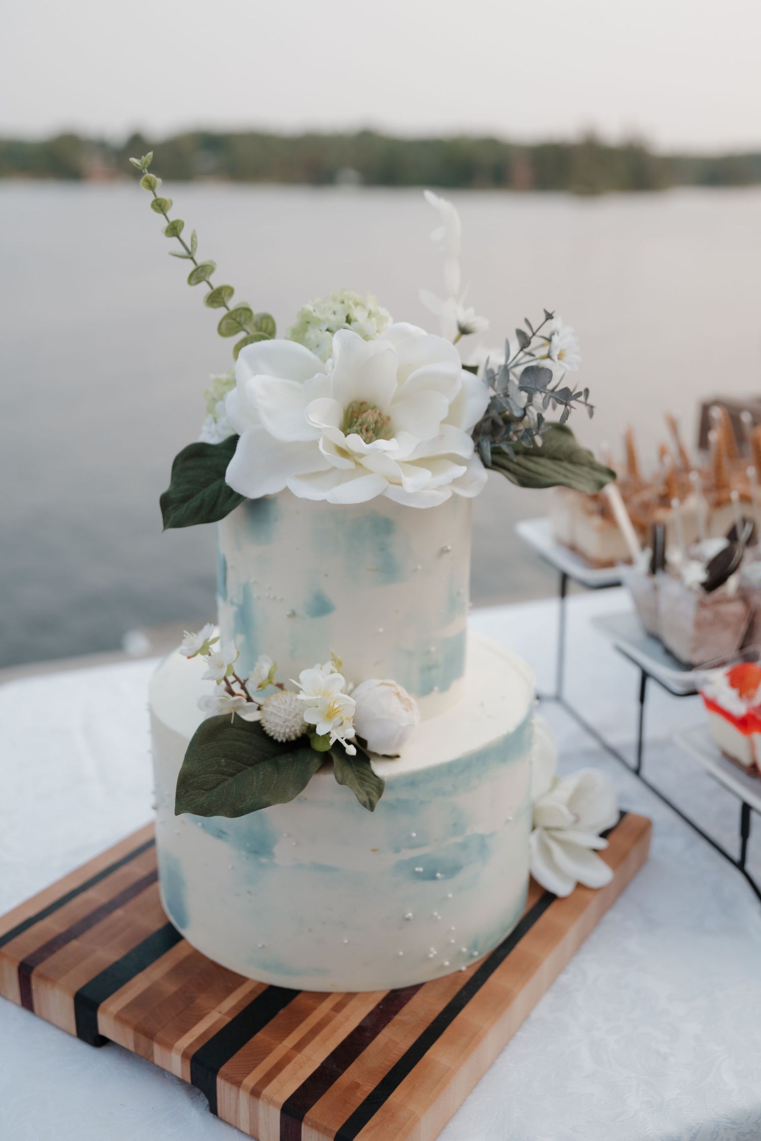 Elegant white wedding cake with light blue watercolor details and fresh floral decorations on a wooden stand.