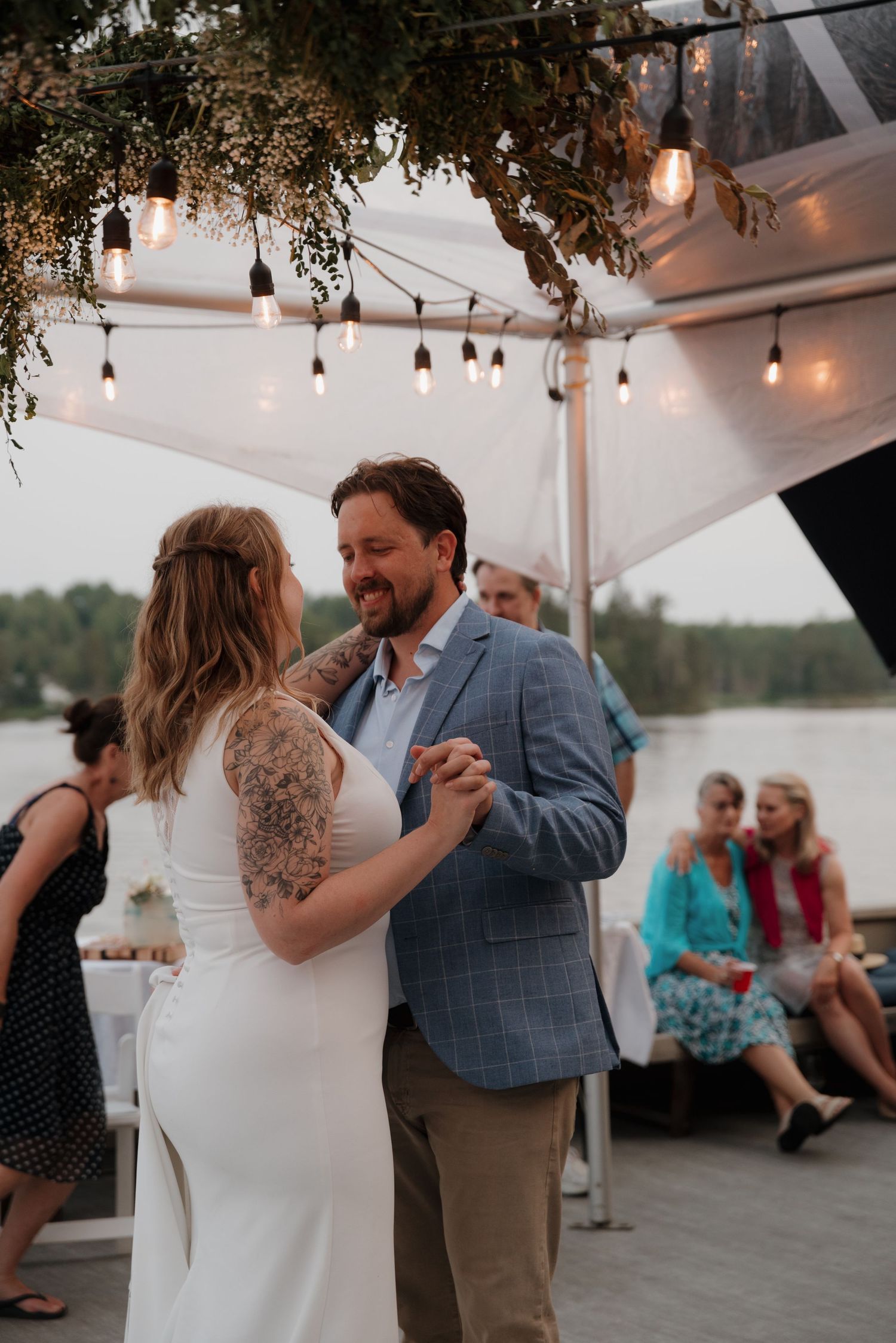 Wedding reception under a white tent with hanging lights and greenery decorations overlooking a lake.