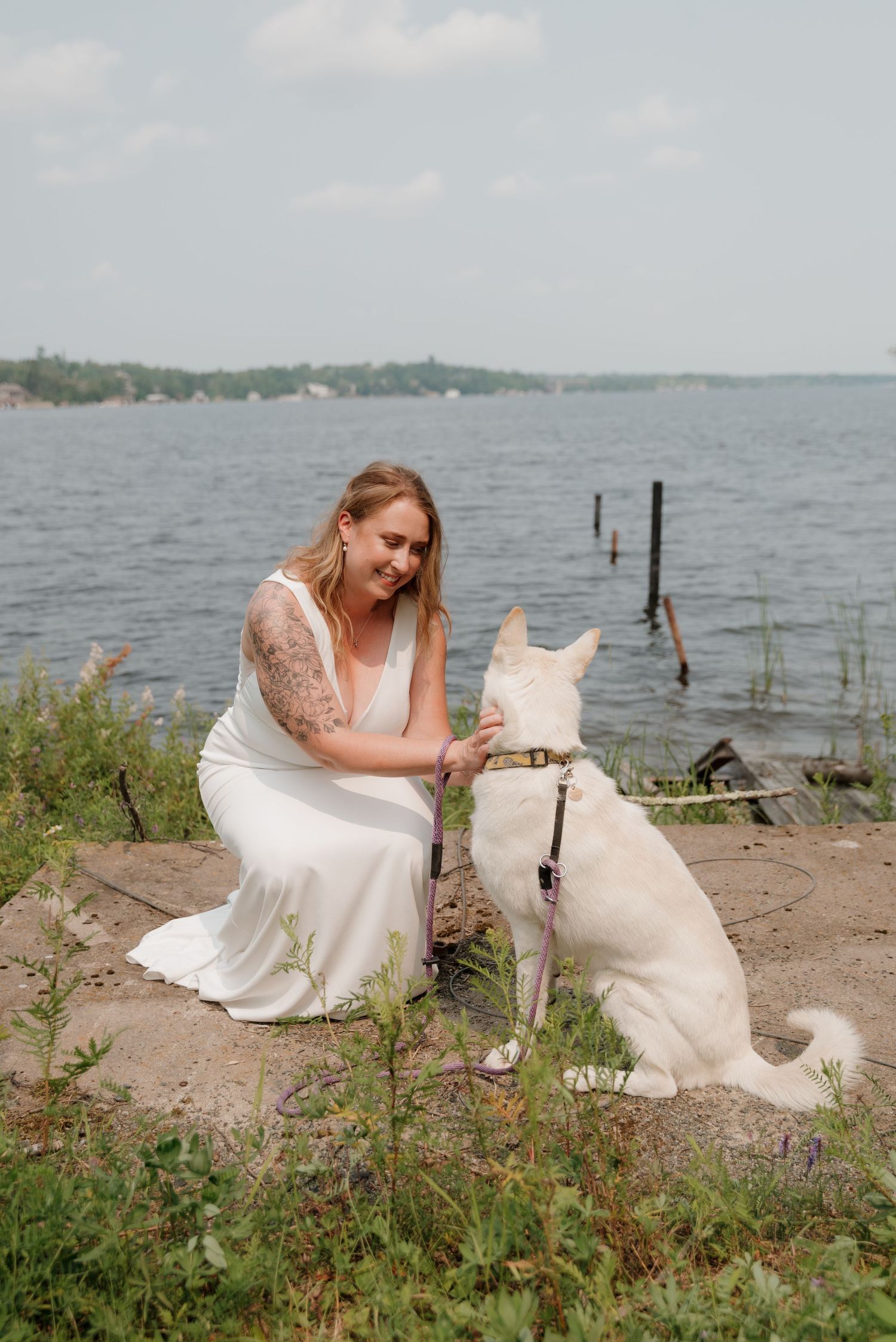 A white dog plays on a rocky shoreline by a lake with boats visible in the background.