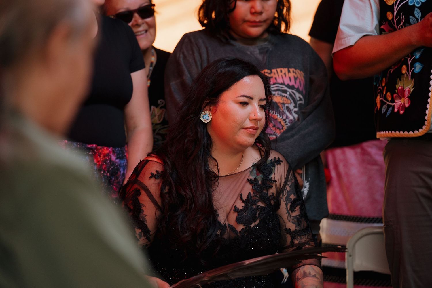 Woman with long dark hair in black lace top sits thoughtfully at an outdoor event.