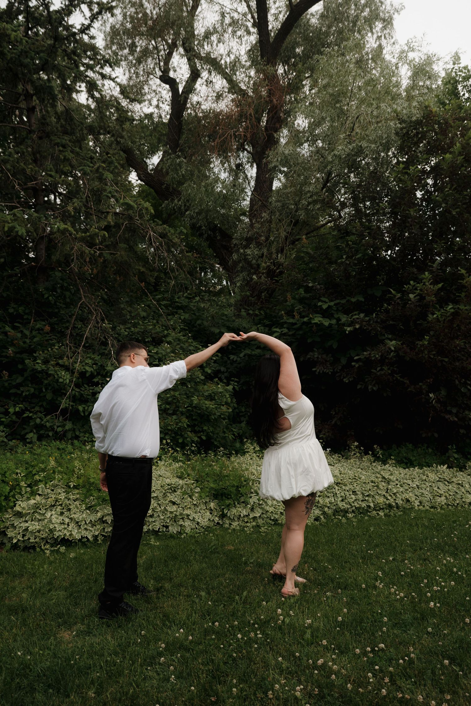 A couple dances playfully in a grassy field surrounded by trees on a summer day.
