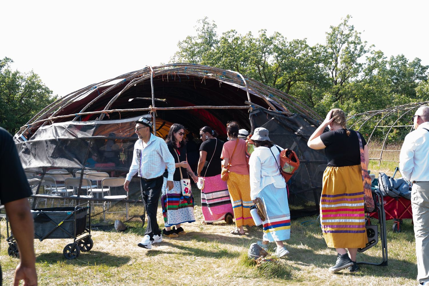 Native American people in traditional dress gather outdoors near a large ceremonial structure on a sunny day.