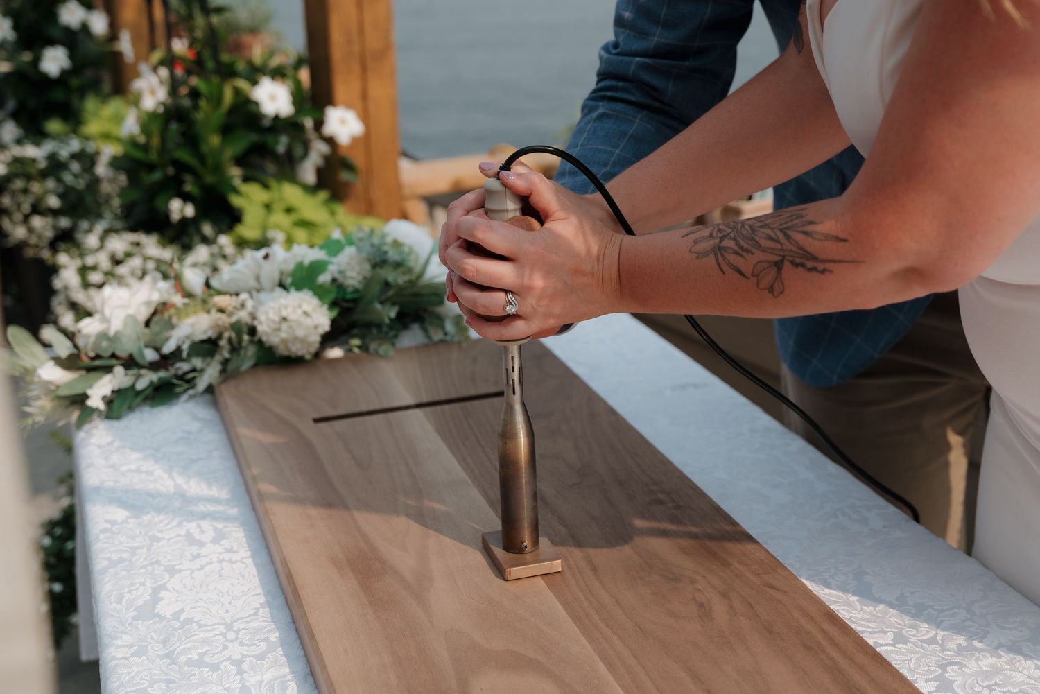 Tattooed hands hold a screwdriver while assembling furniture near floral decorations on a white surface.
