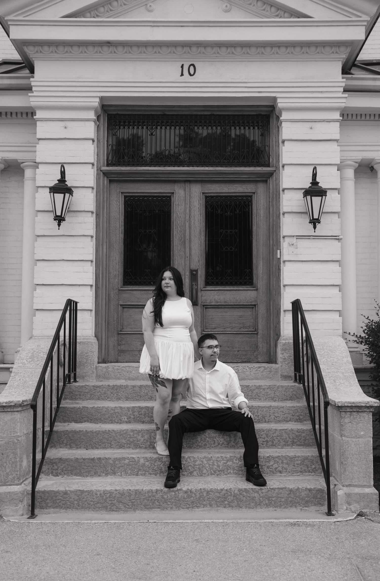 A black and white photo of a couple posing on steps of a classic building entrance with lanterns.