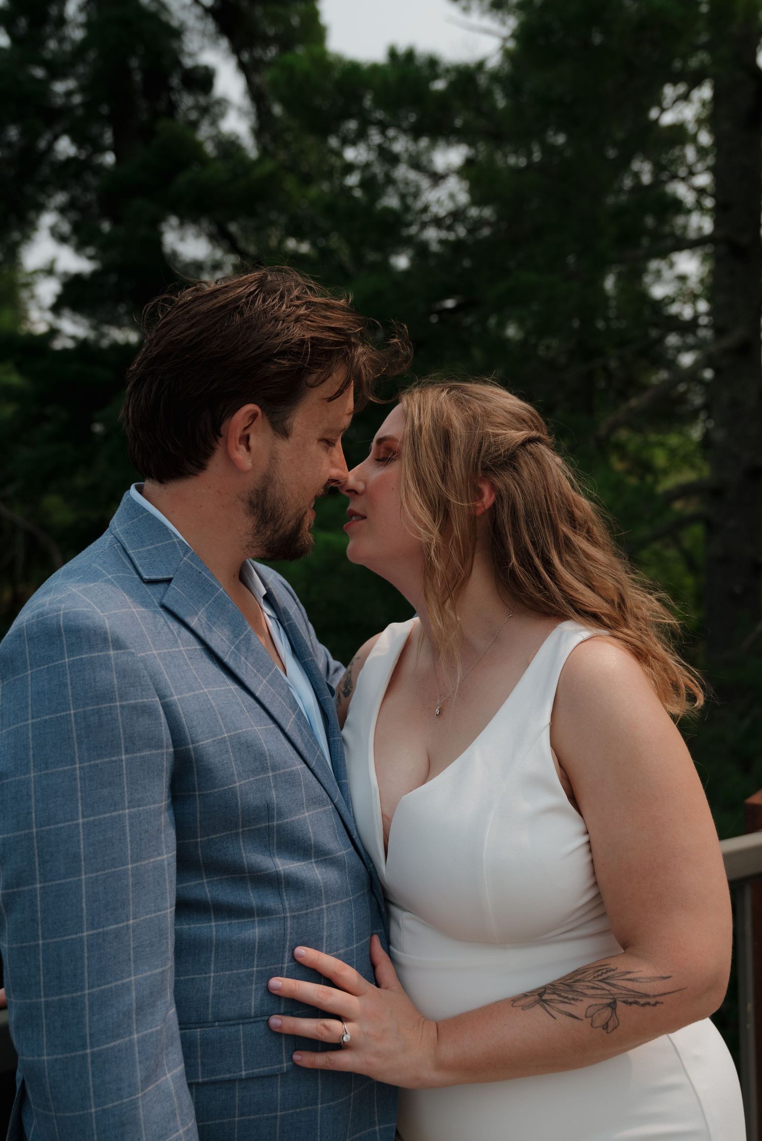 A romantic wedding couple embraces outdoors, with the groom wearing a light blue suit and the bride in a white dress.