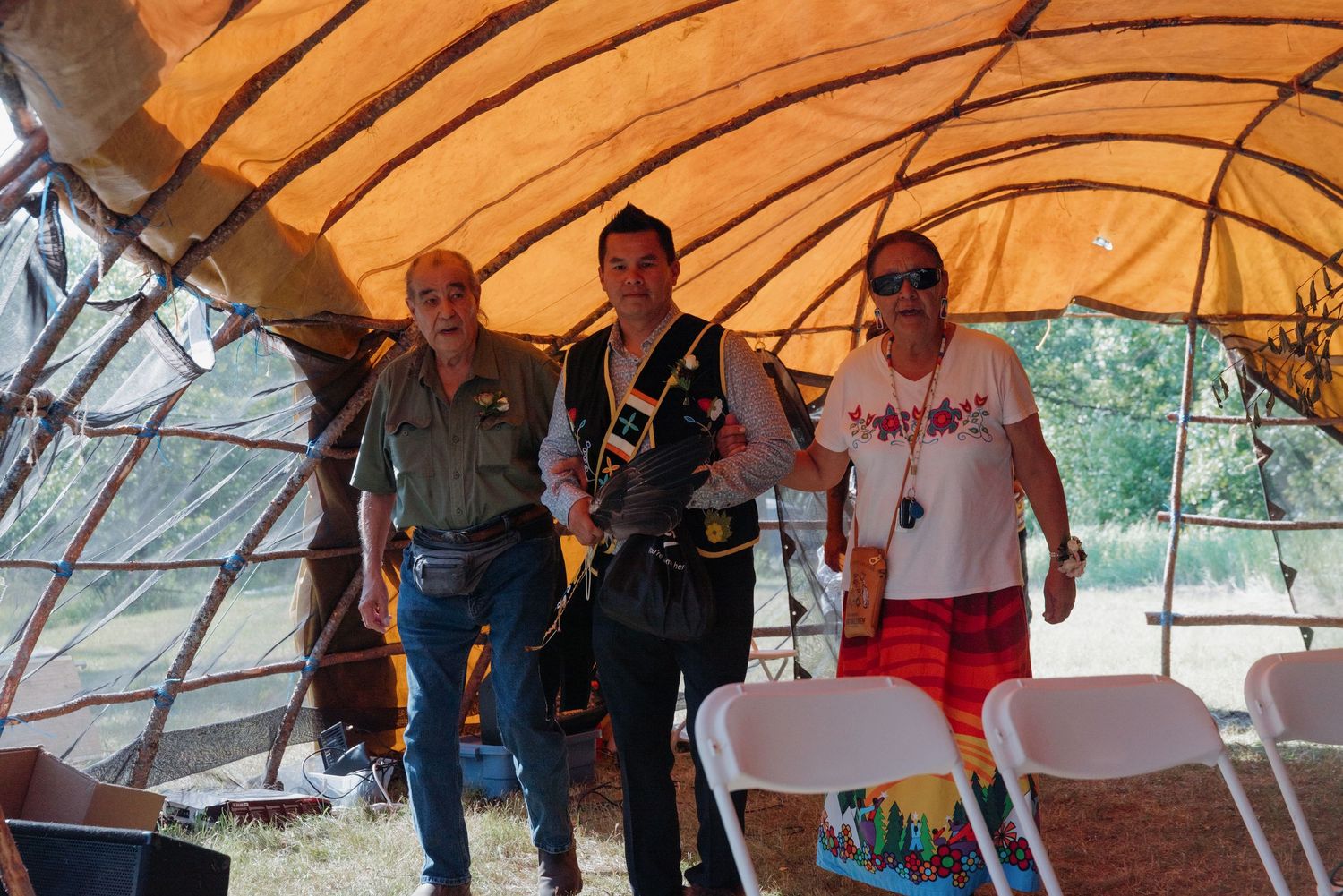 Three people standing inside a traditional Native American arbor structure with orange hide walls and white folding chairs.