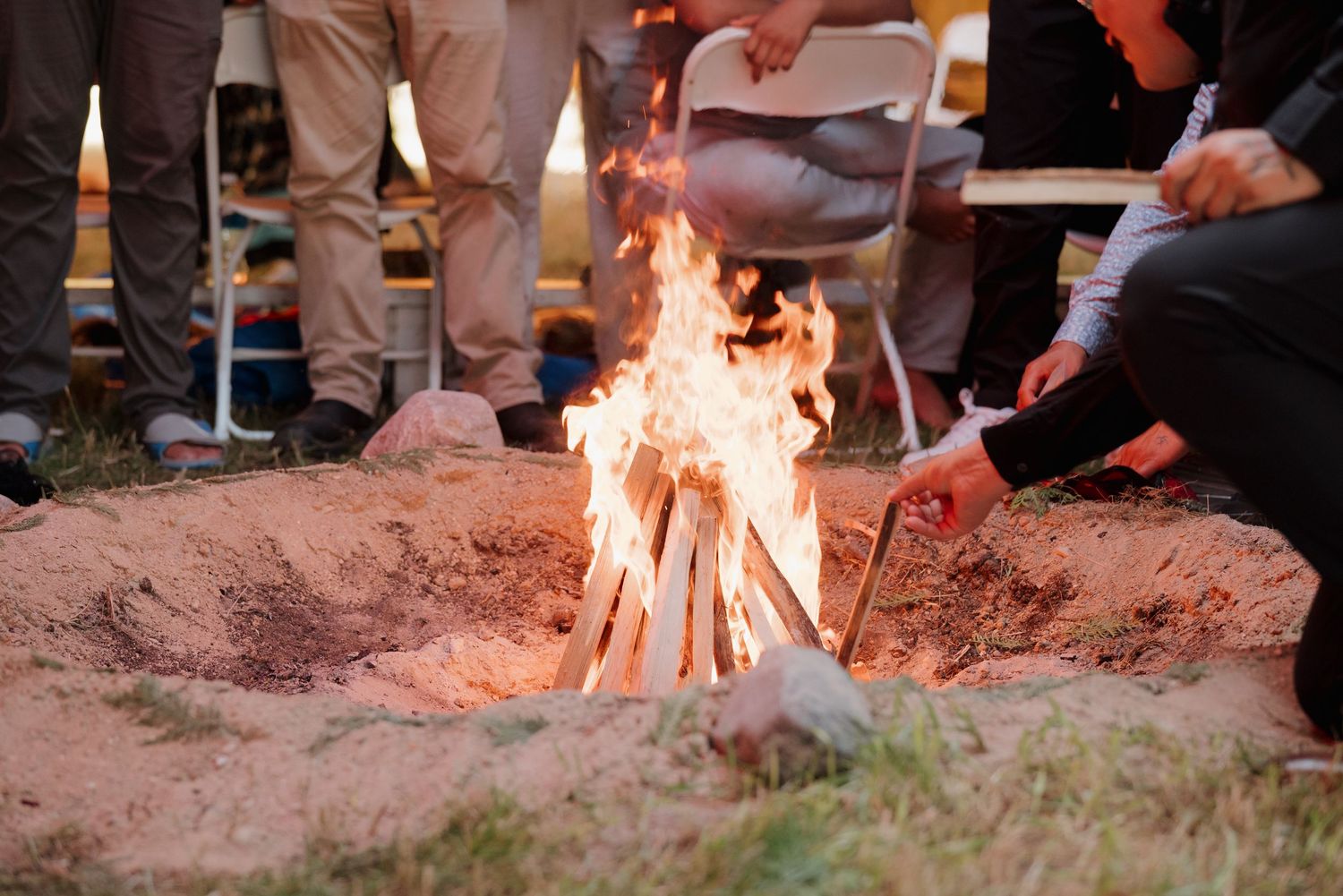 Group of people gathered around a glowing campfire in a sandy pit outdoors at dusk.