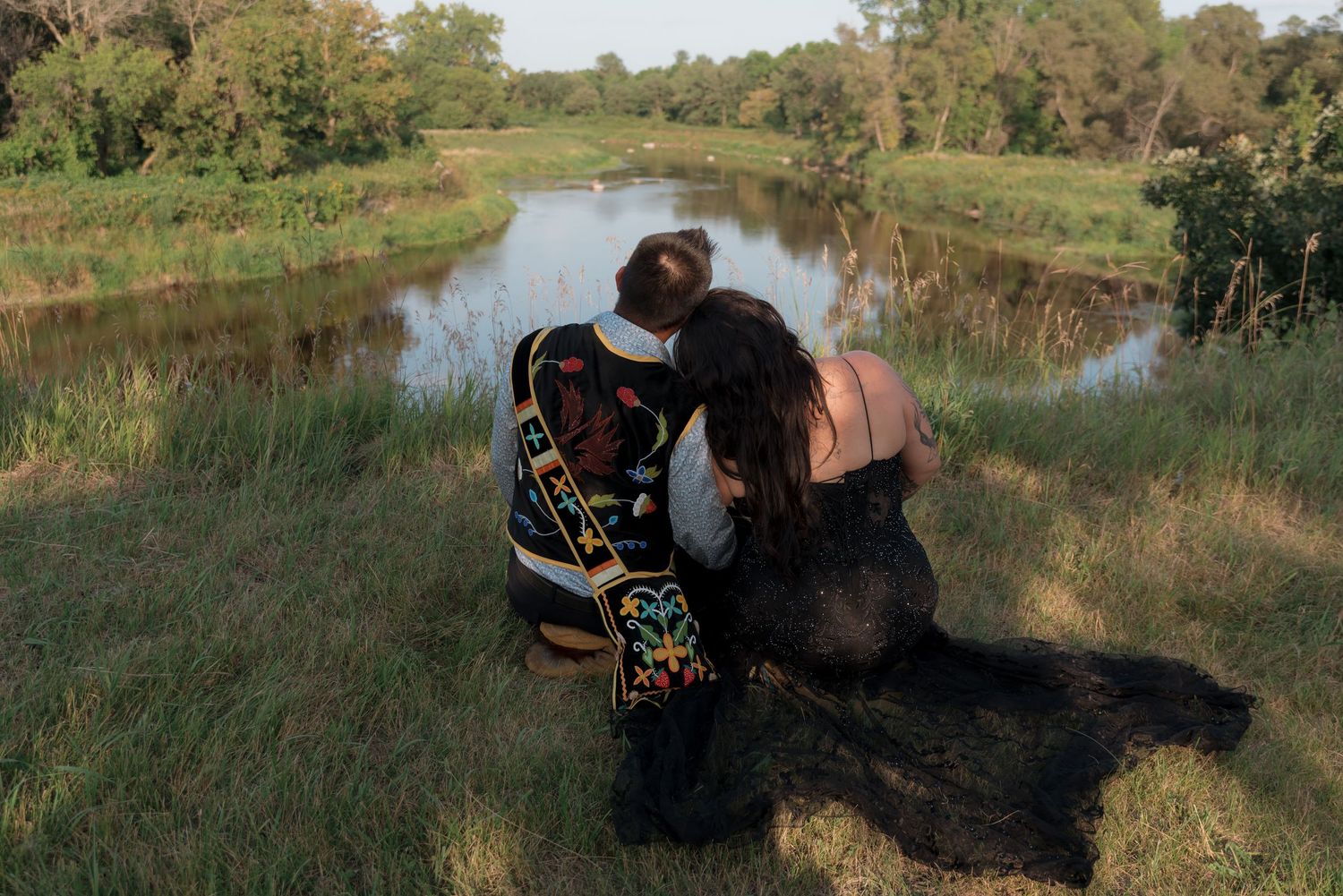 Two people sitting together on grass overlooking a peaceful river surrounded by lush green trees.