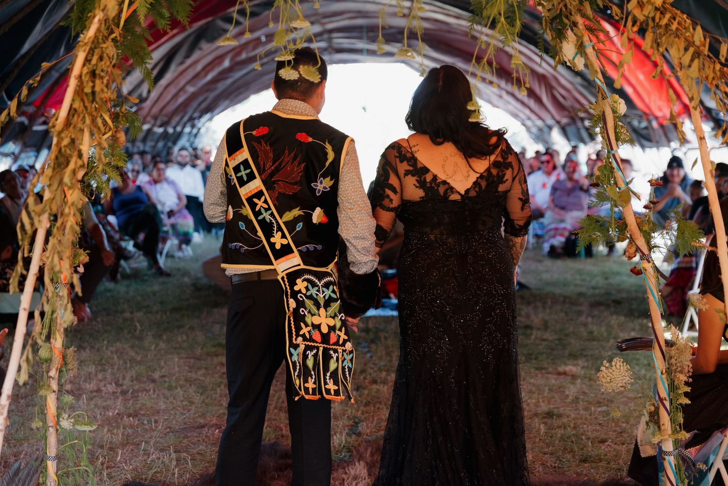 Couple stands under floral arch in traditional Indigenous wedding ceremony, groom wearing decorative cultural sash and regalia.