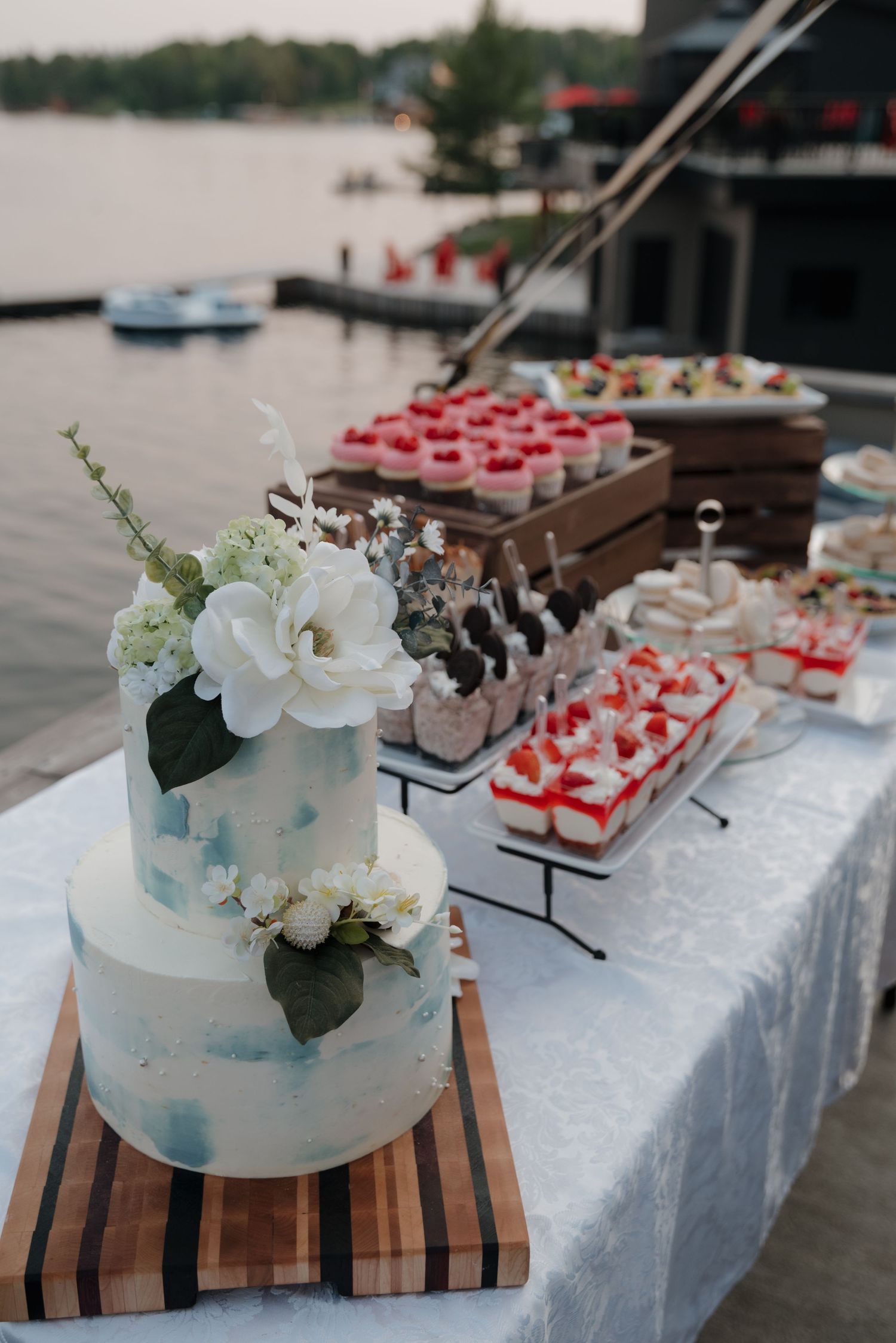 Elegant wedding dessert table with white cake and colorful petit fours displayed on wooden boards by the waterfront.