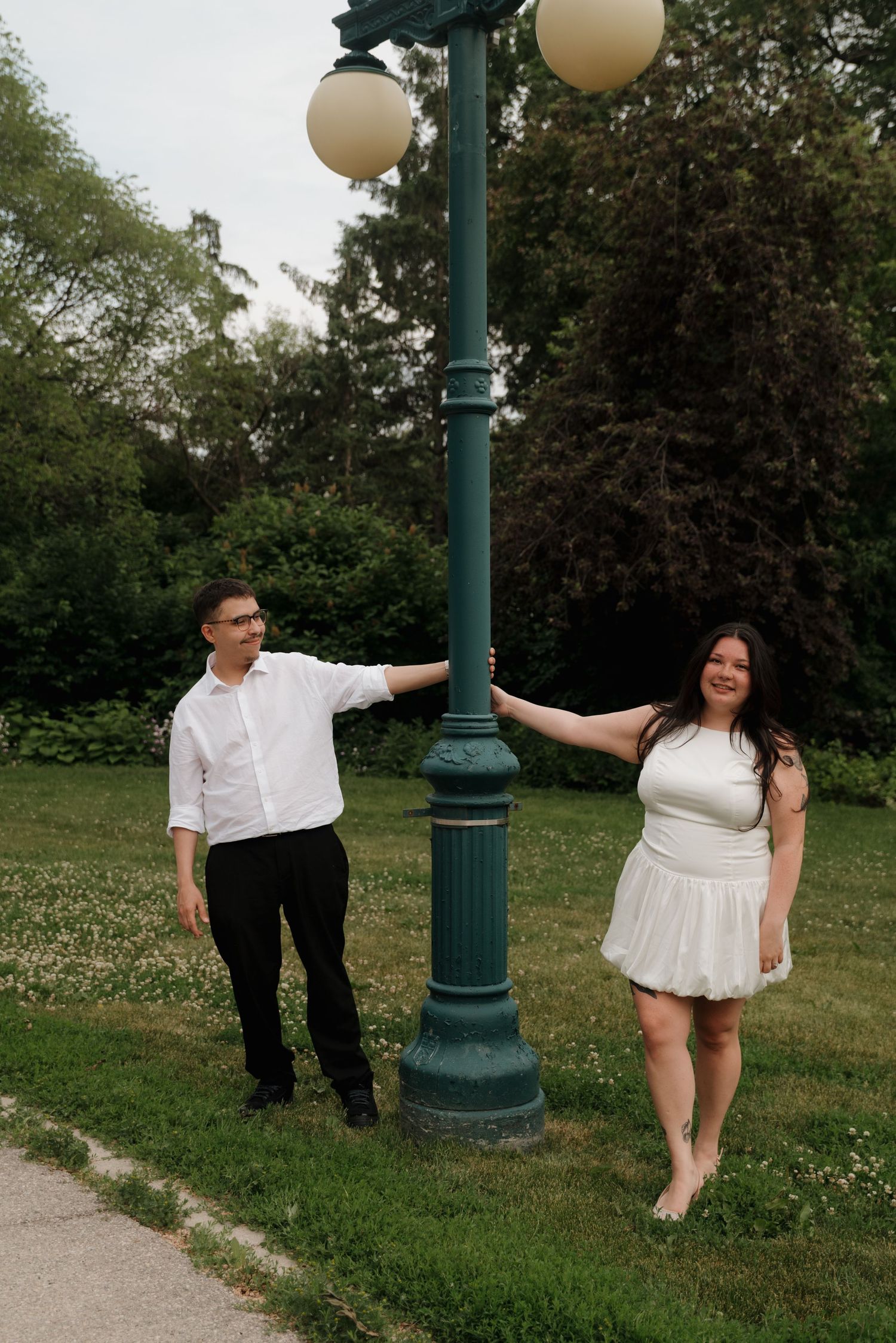 A couple poses playfully by a vintage-style green lamp post in a park setting with grass and trees.