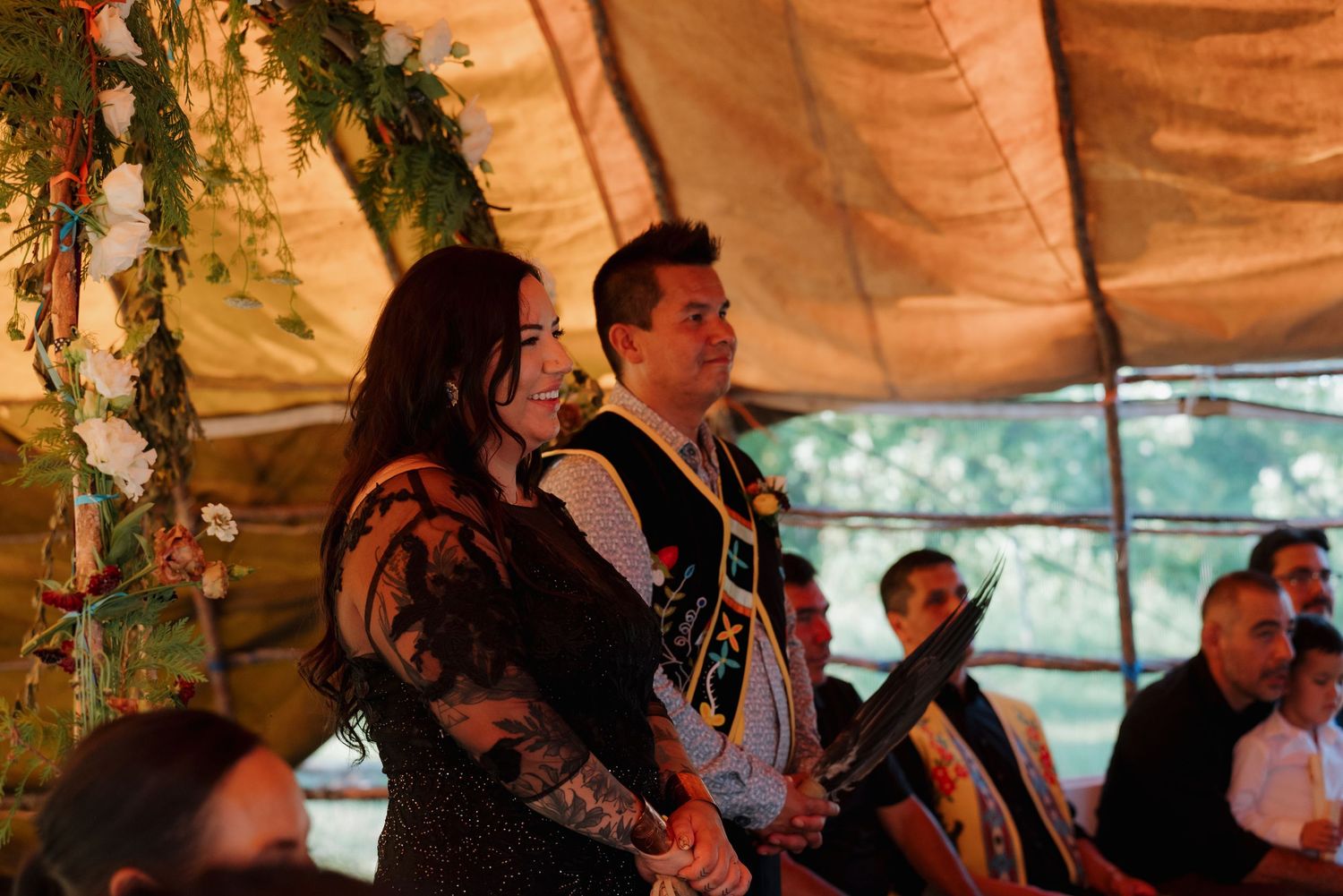 A couple smiles during an outdoor ceremony under a decorated tent with floral arrangements.
