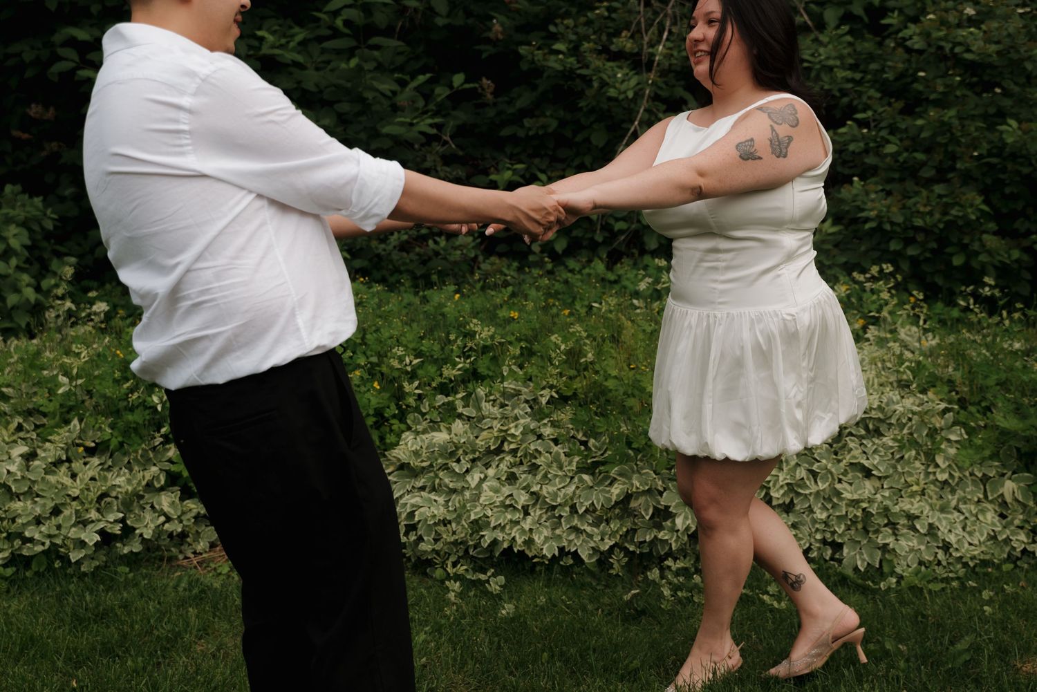 A couple in white outfits dance together on grass in a romantic outdoor setting.