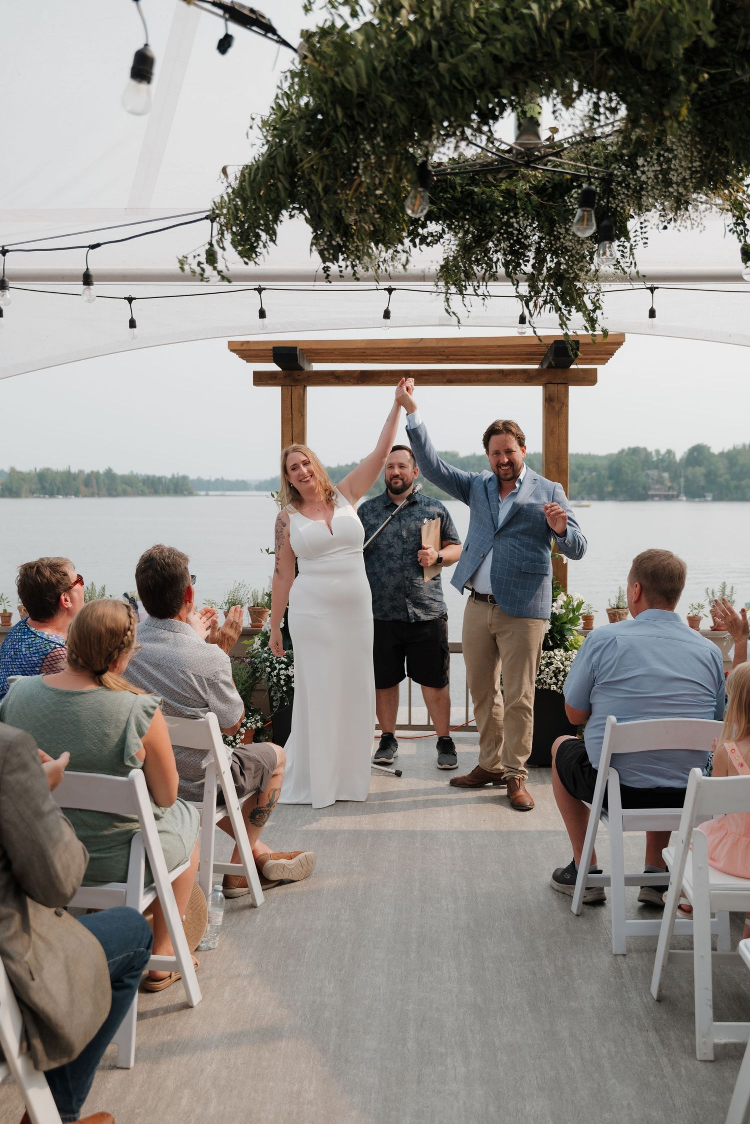 Guests seated at an outdoor waterfront wedding ceremony under string lights and trees.