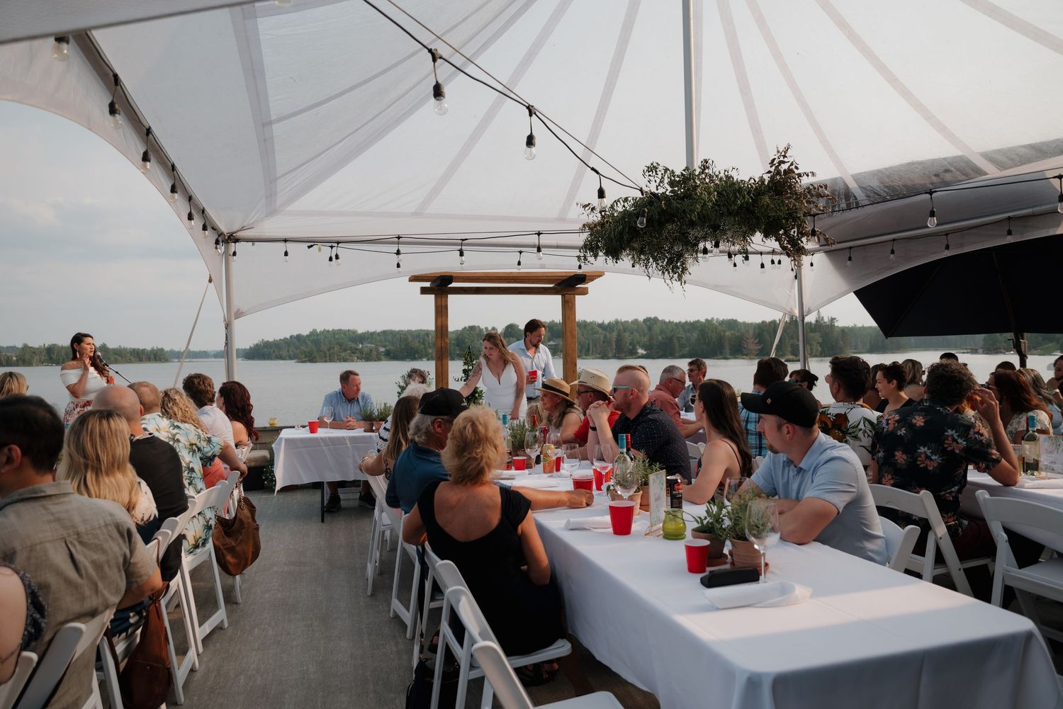 Wedding reception setup under white tent with string lights and waterfront views.