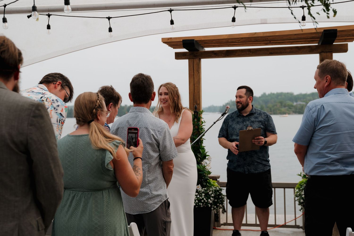 Small group gathered at waterfront wedding ceremony with wooden arch and string lights.