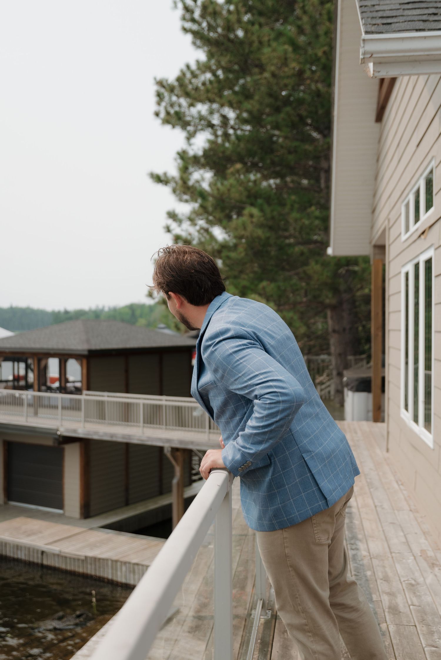 Figure leaning on white deck railing overlooking lake with pine trees in background.