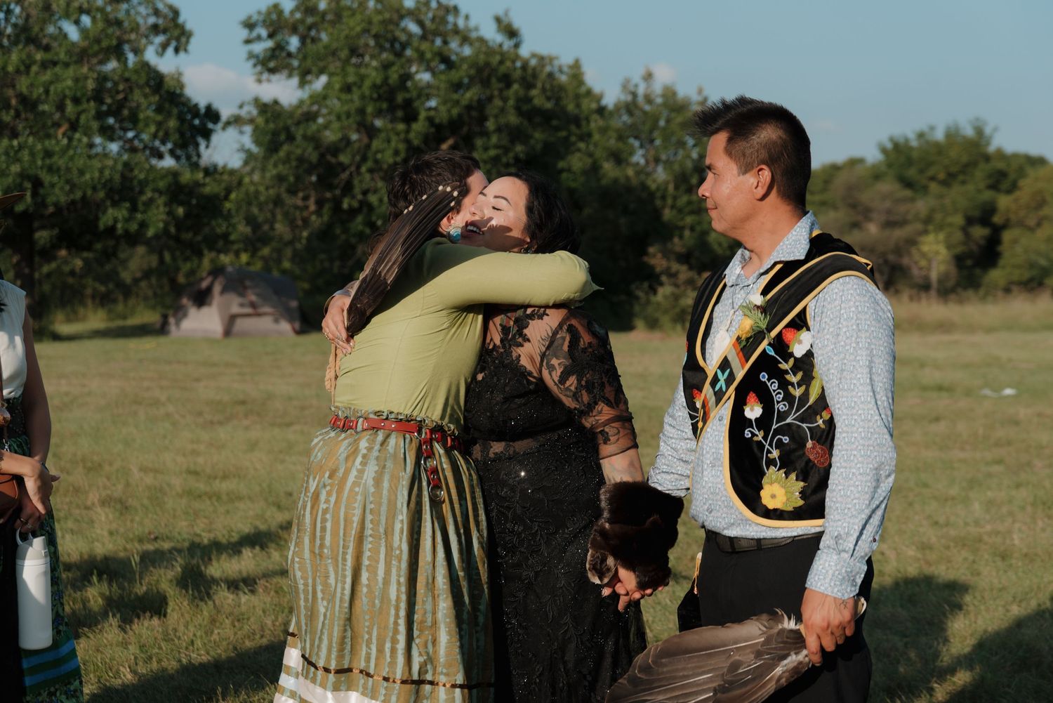 Two women in traditional Native American clothing share a warm embrace while a man stands nearby outdoors.