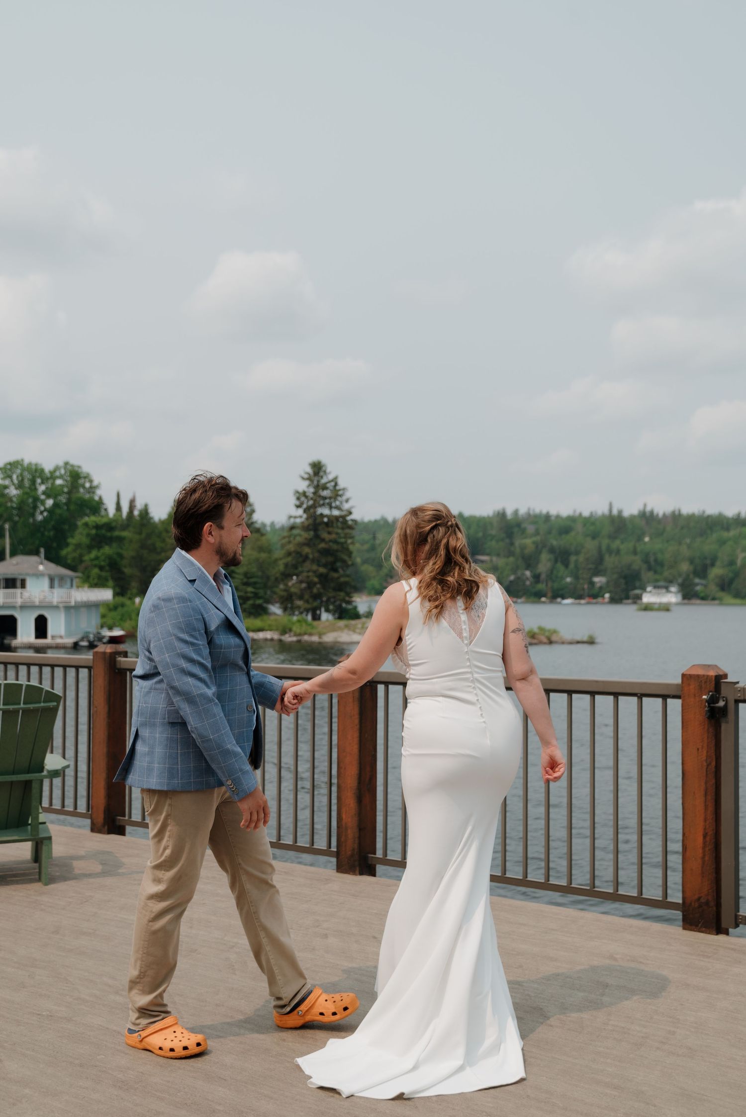 A couple in wedding attire playfully poses on a lakeside dock with trees and water in the background.