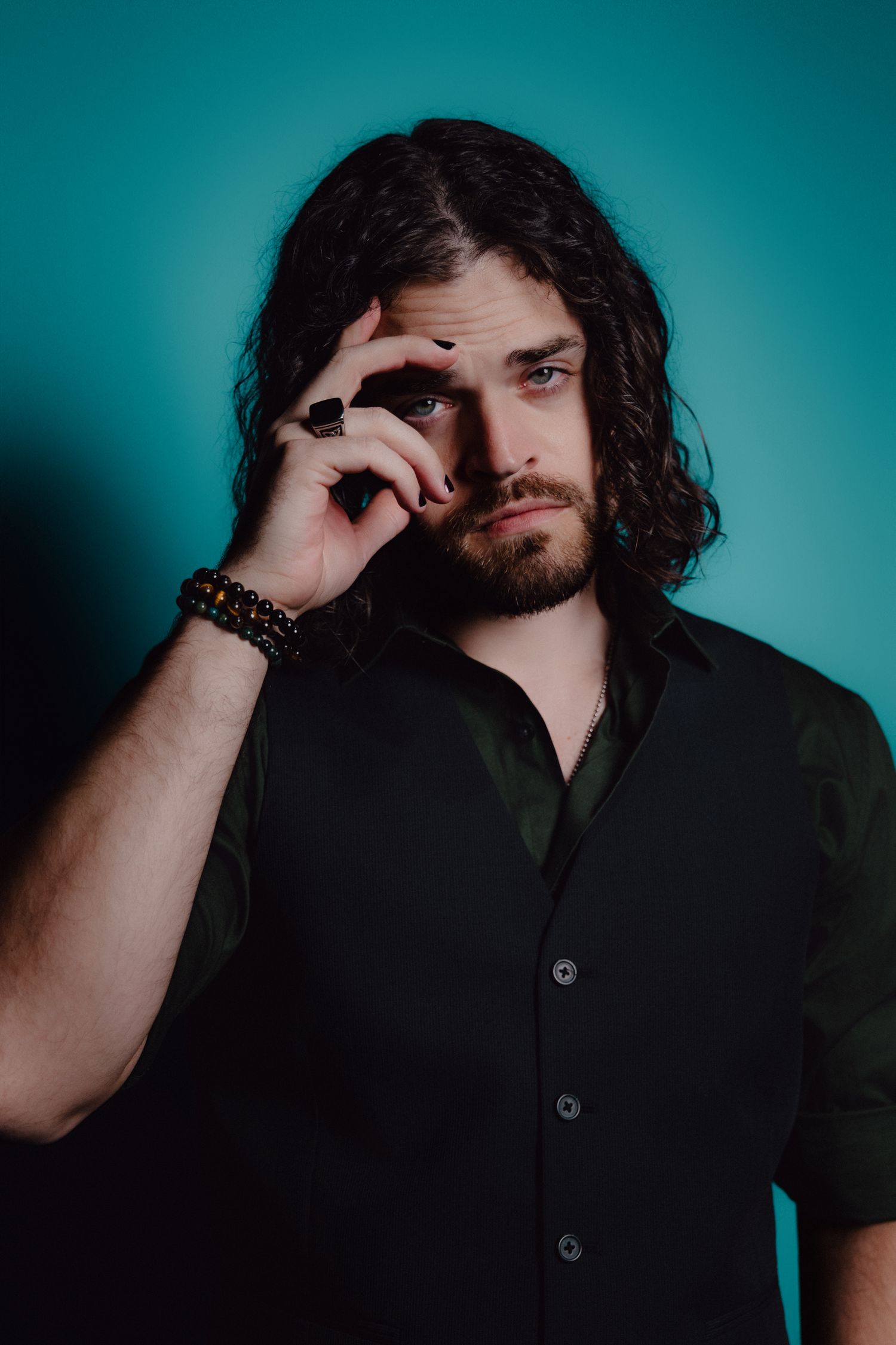 Young man with long dark hair and beard in black shirt poses against teal background.