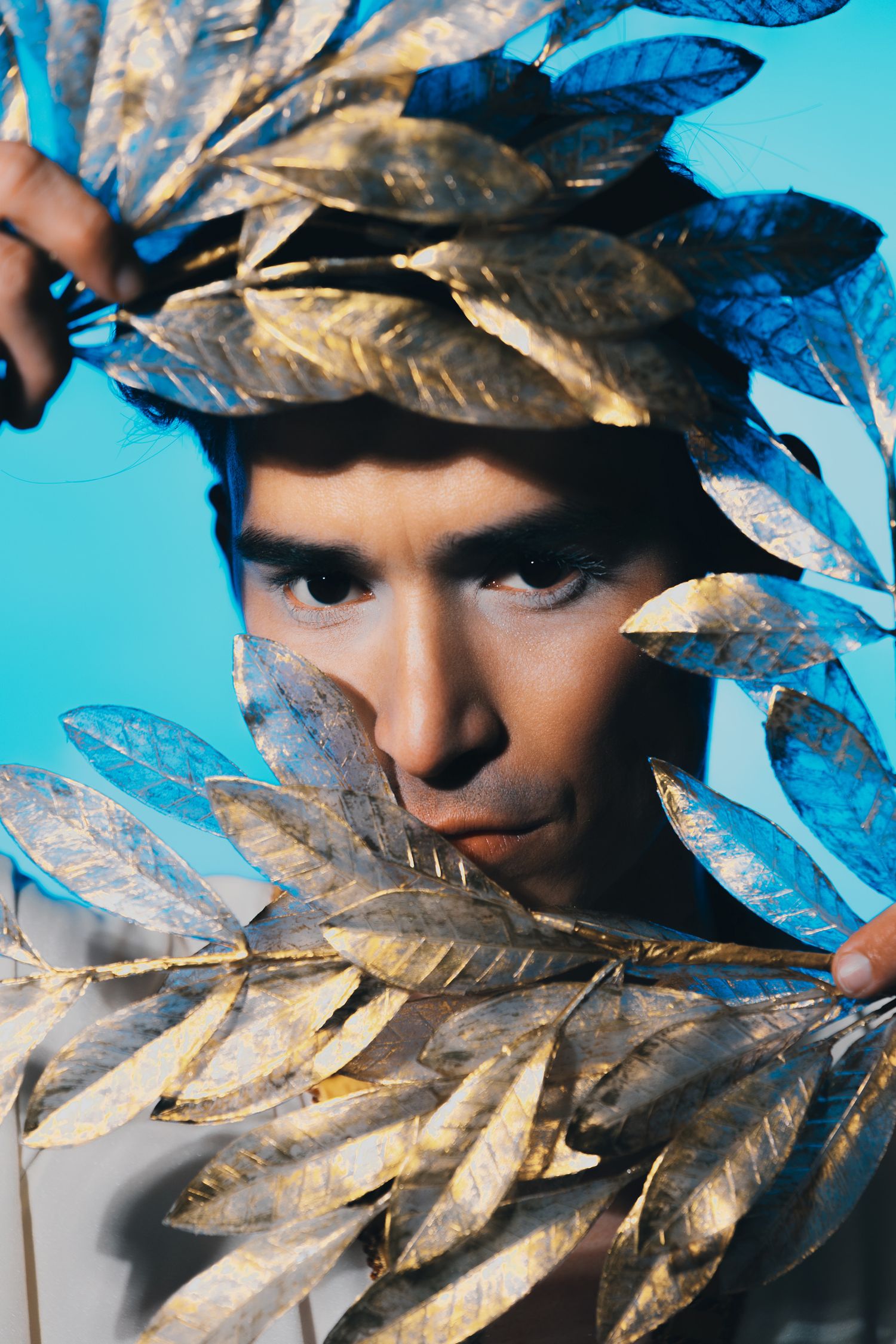 Man surrounded by golden and blue leaves against a bright blue background, wearing a laurel crown in an artistic portrait.