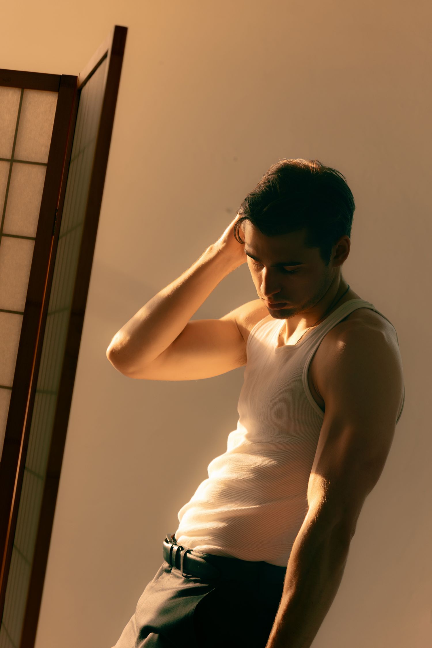 Muscular man in white tank top leaning against wall with hand on head in dramatic warm lighting.