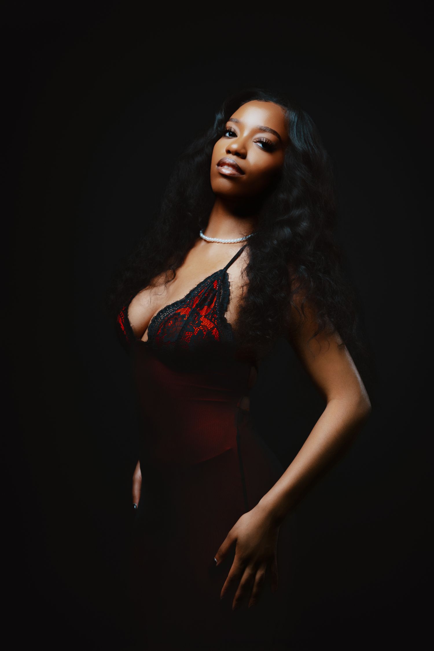 A young Black woman in red and black lingerie poses confidently against a dark background with long curly hair.