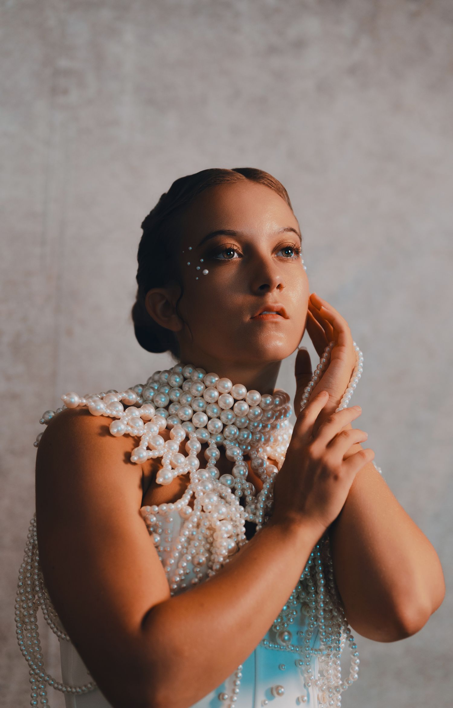 Woman in pearl-embellished top with pearl face gems poses gracefully against gray backdrop.