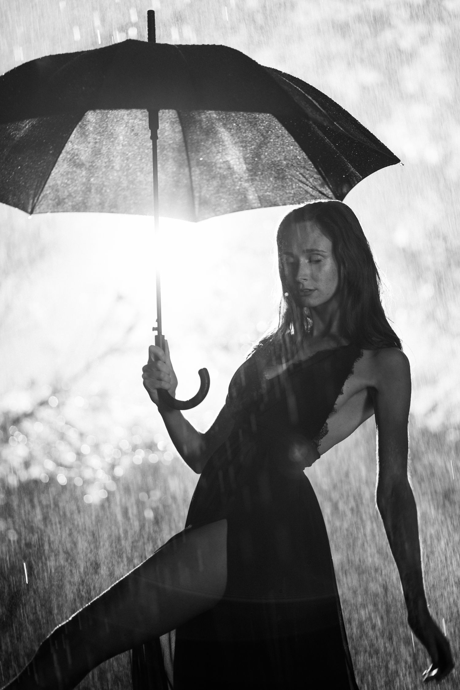 Black and white photo of a woman in a black dress holding an umbrella in the rain with backlit bokeh background.