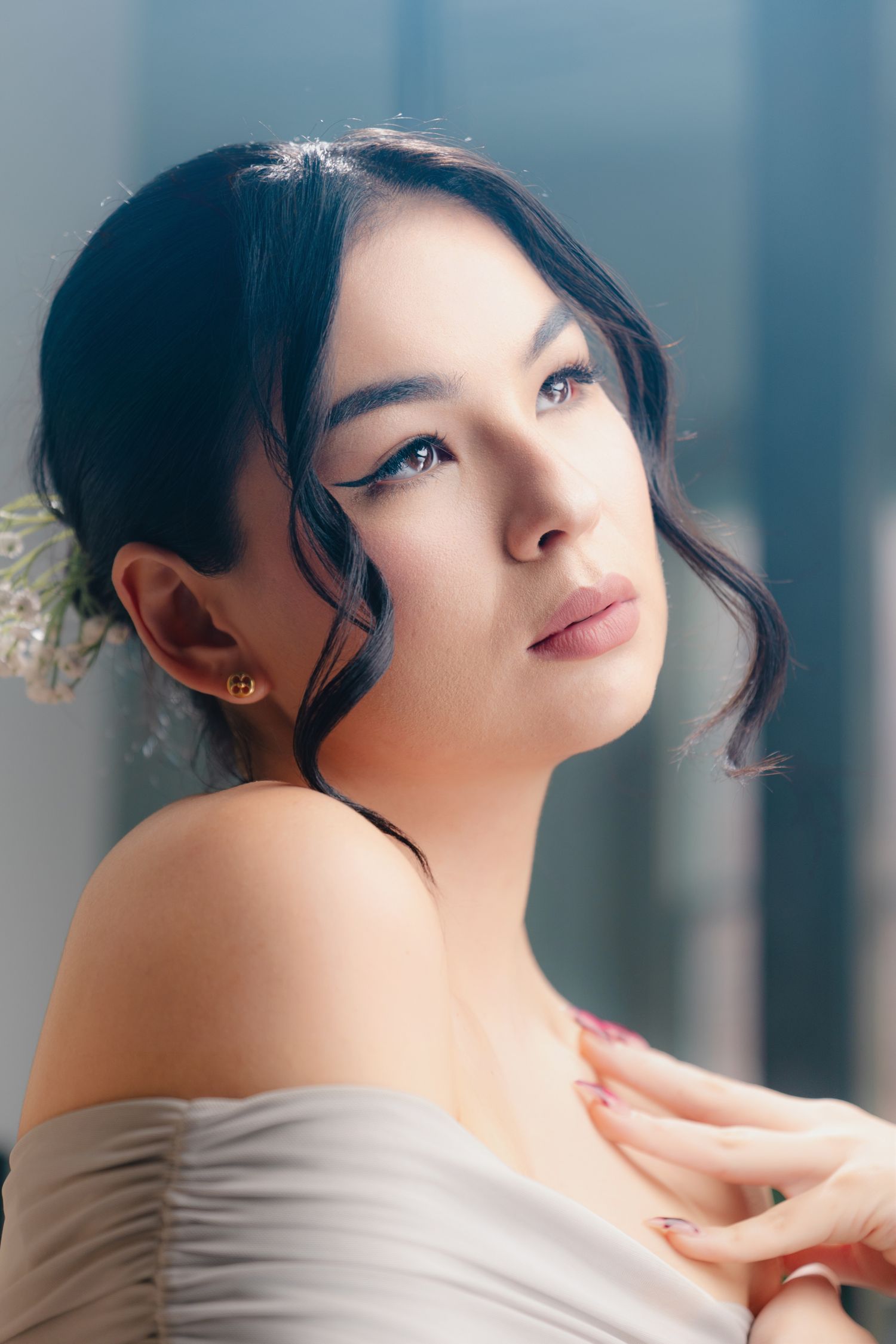 Young Asian woman with elegant updo, floral hair accessory, winged eyeliner, and off-shoulder white dress poses gracefully.