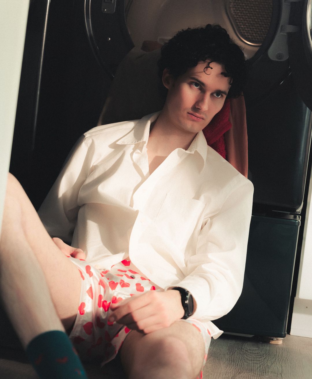 Young man in white shirt and heart-print shorts sitting casually on floor, wearing teal socks and a watch.