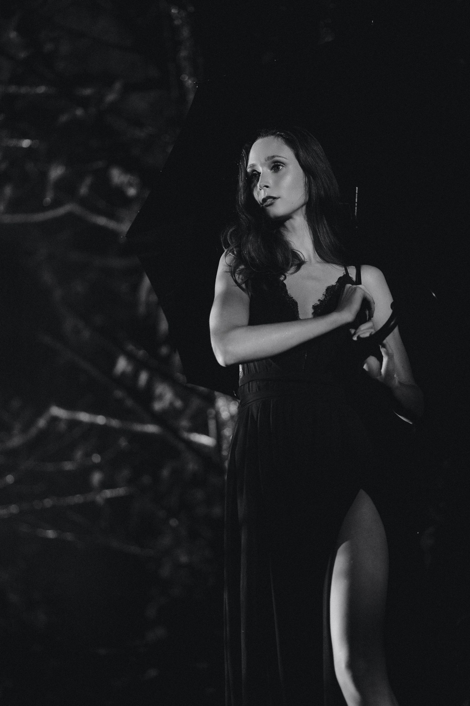 Black and white photo of a woman in an elegant dark gown standing in a moody, shadowy outdoor setting.