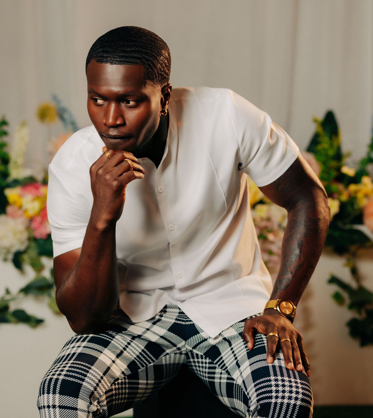 A stylish young Black man in a white shirt and plaid pants poses thoughtfully against a floral background.