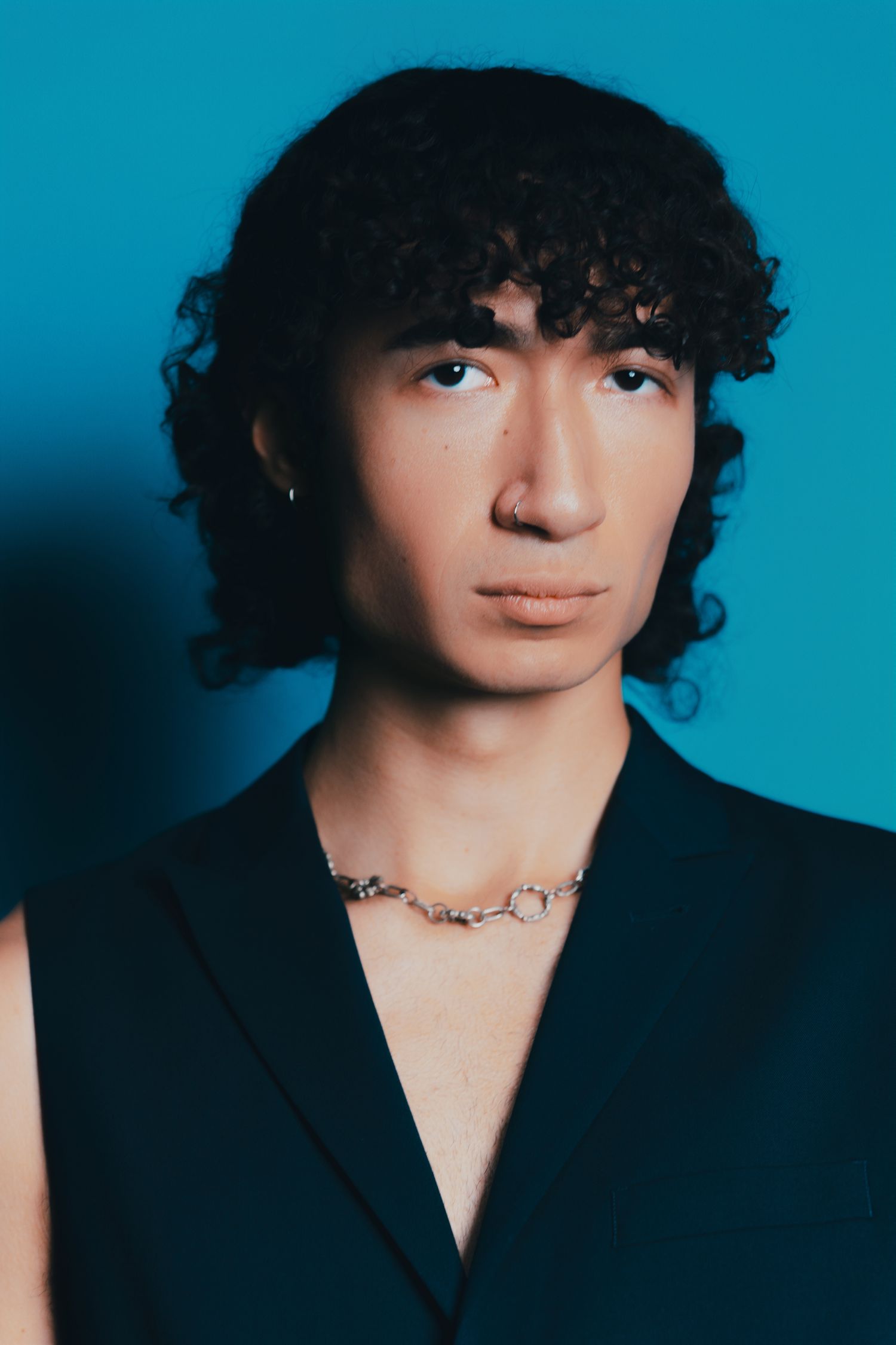 Young person with curly dark hair wearing a black vest and chain necklace poses against a blue background.