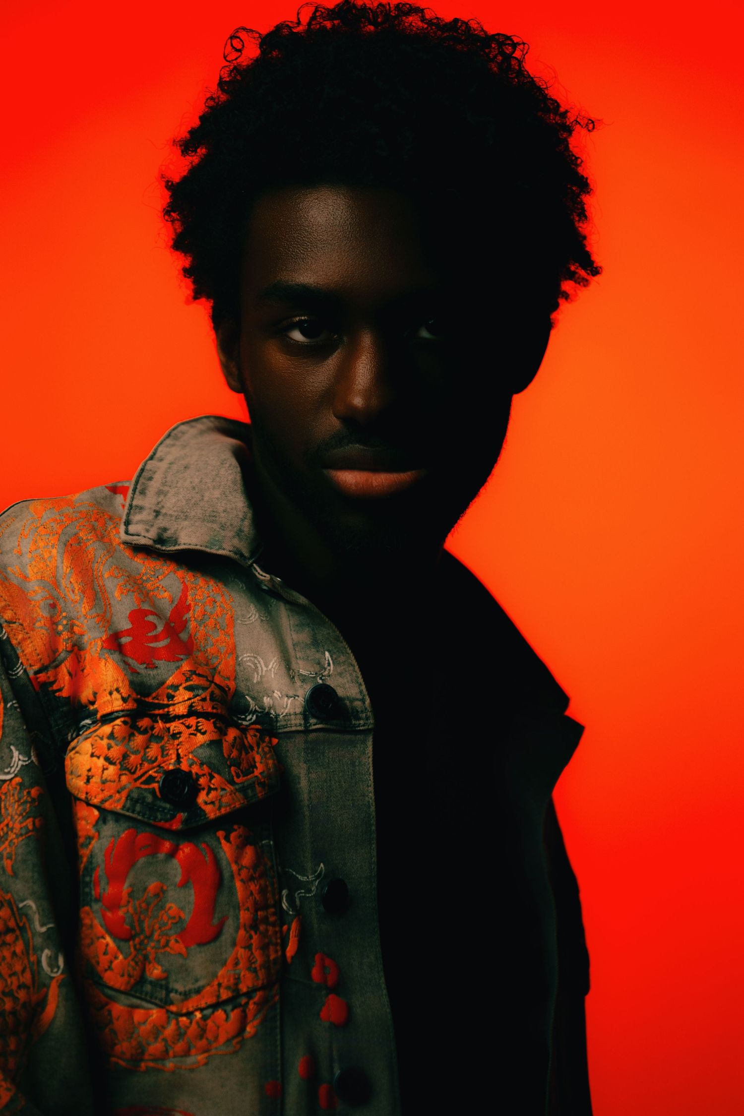 Young Black man in colorful patterned jacket poses against vibrant red background in dramatic studio portrait.