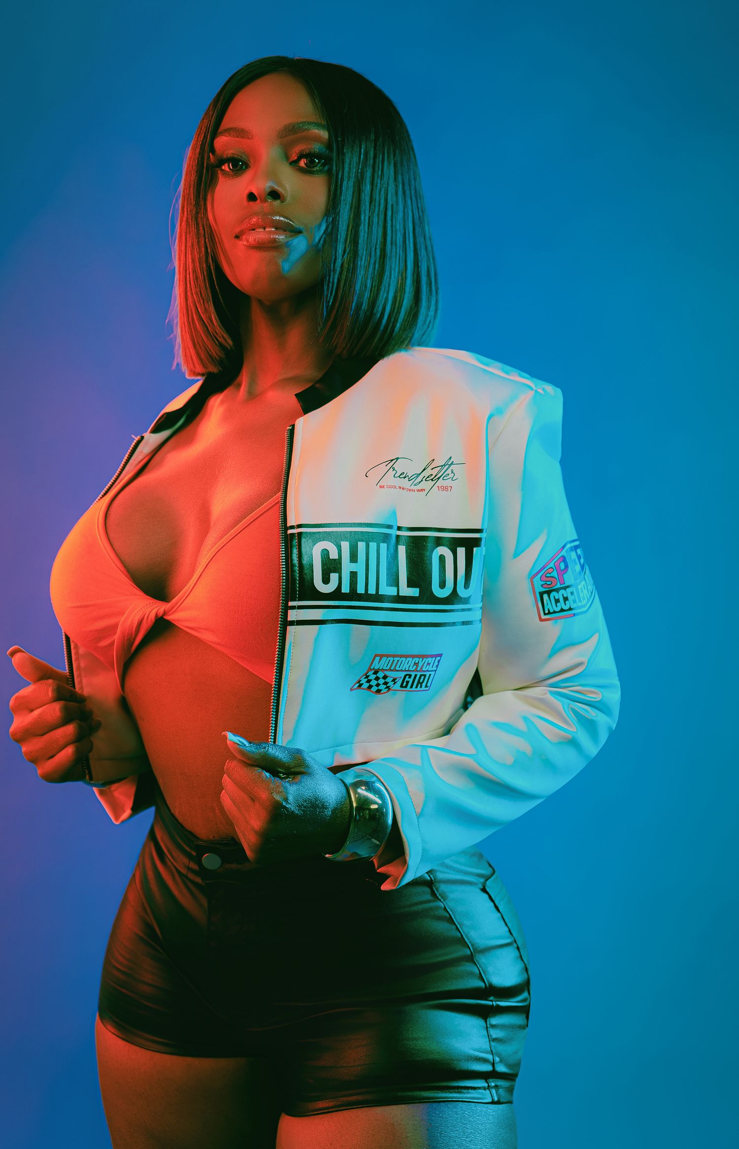 Woman in white 'Chill Out' jacket and black shorts poses confidently under vibrant blue and red studio lighting.