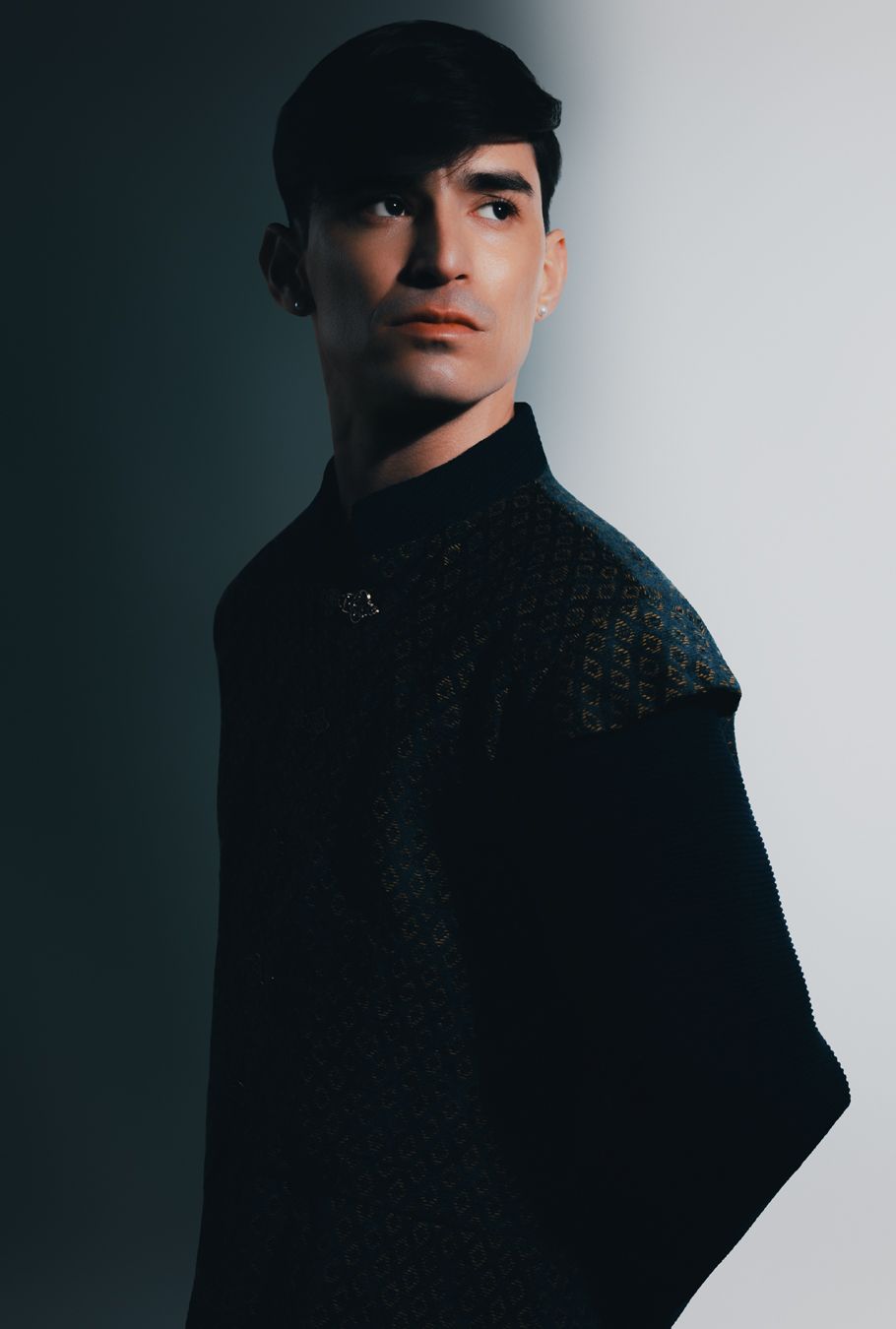 Young man in dark cape with patterned shoulder detail poses dramatically against a two-tone gray background.