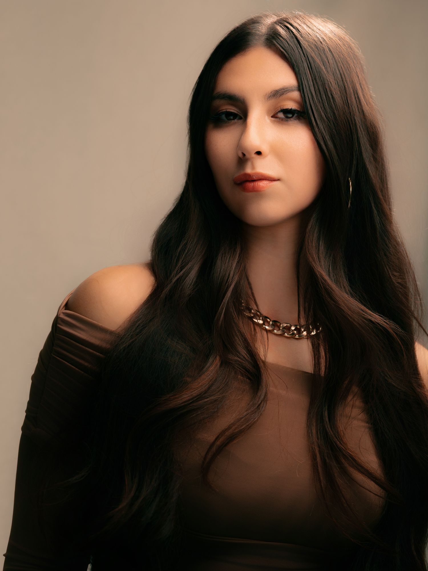 Young woman with long dark hair wearing a black off-shoulder top and gold chain necklace poses against a gray background.
