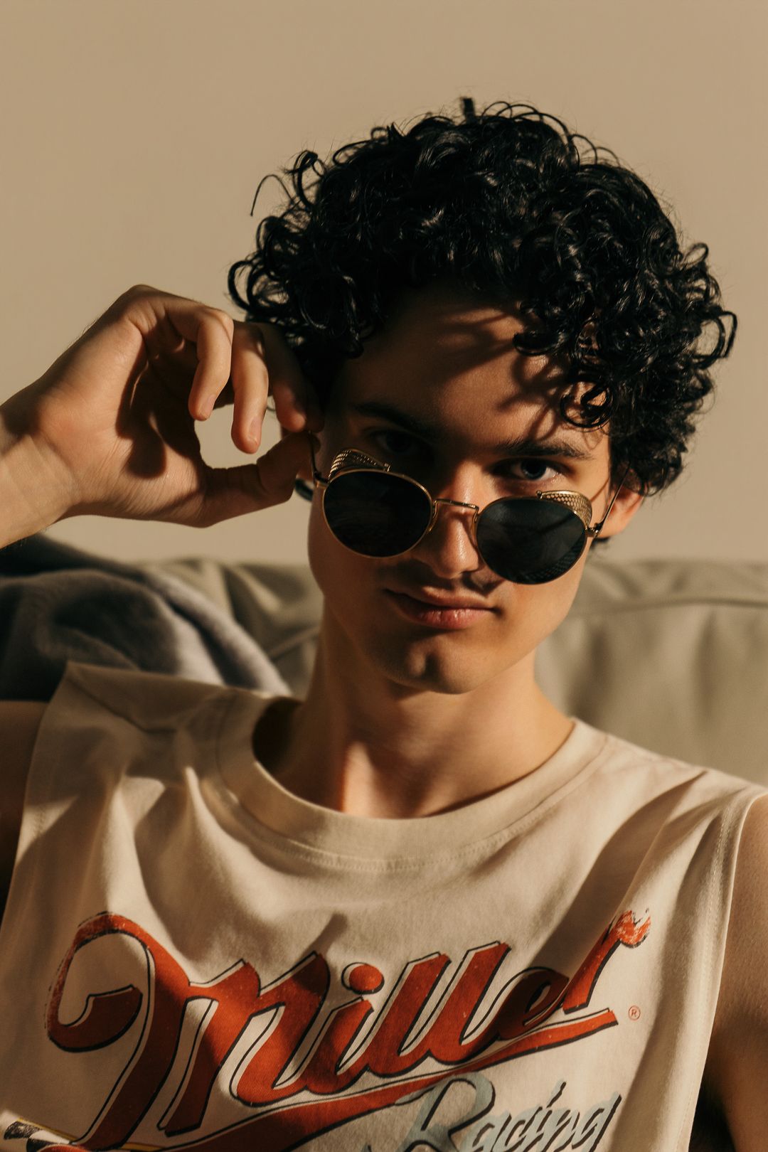 Young person with curly hair wearing sunglasses and a Miller t-shirt posing confidently in warm light.