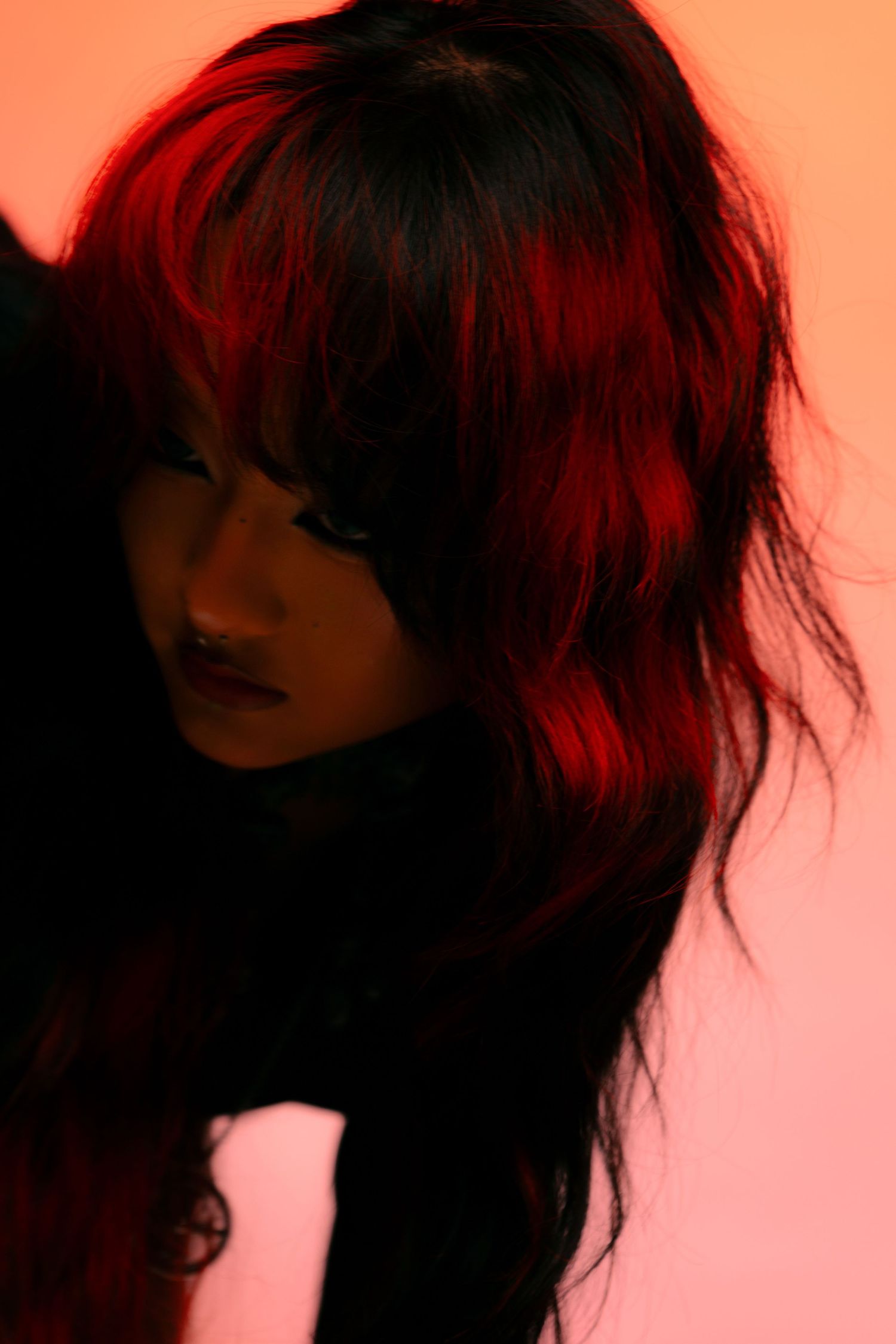 A woman with dramatic red and black hair poses under moody red lighting against a pink background.