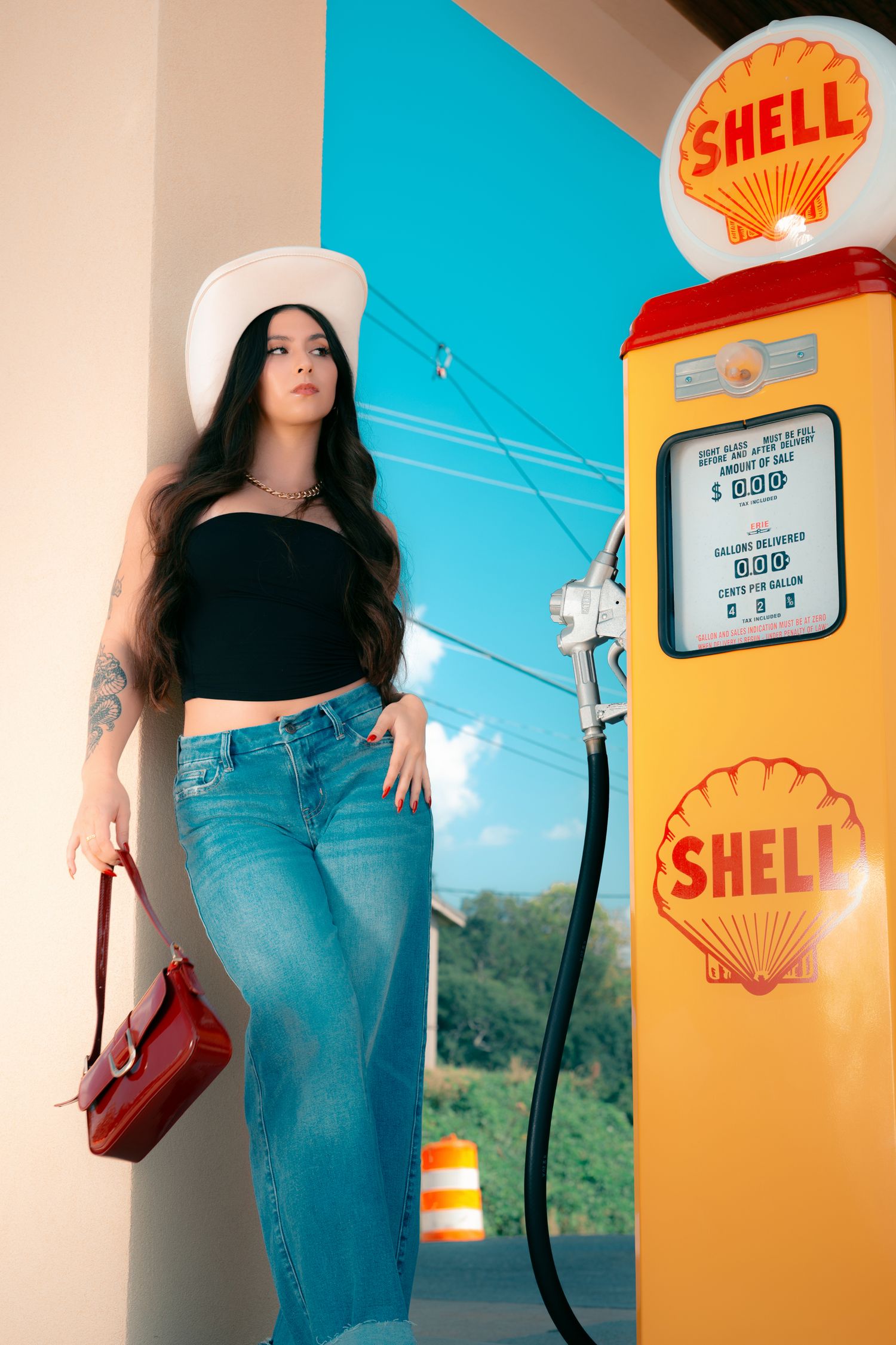 Woman in black crop top, jeans, and white cowboy hat poses beside a vintage yellow Shell gas pump.
