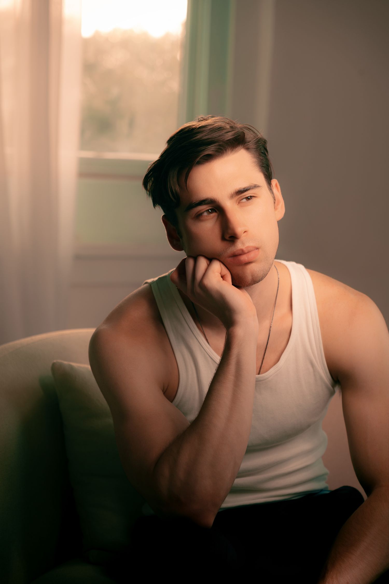 Young man in white tank top sitting pensively by a sunlit window with warm golden light.