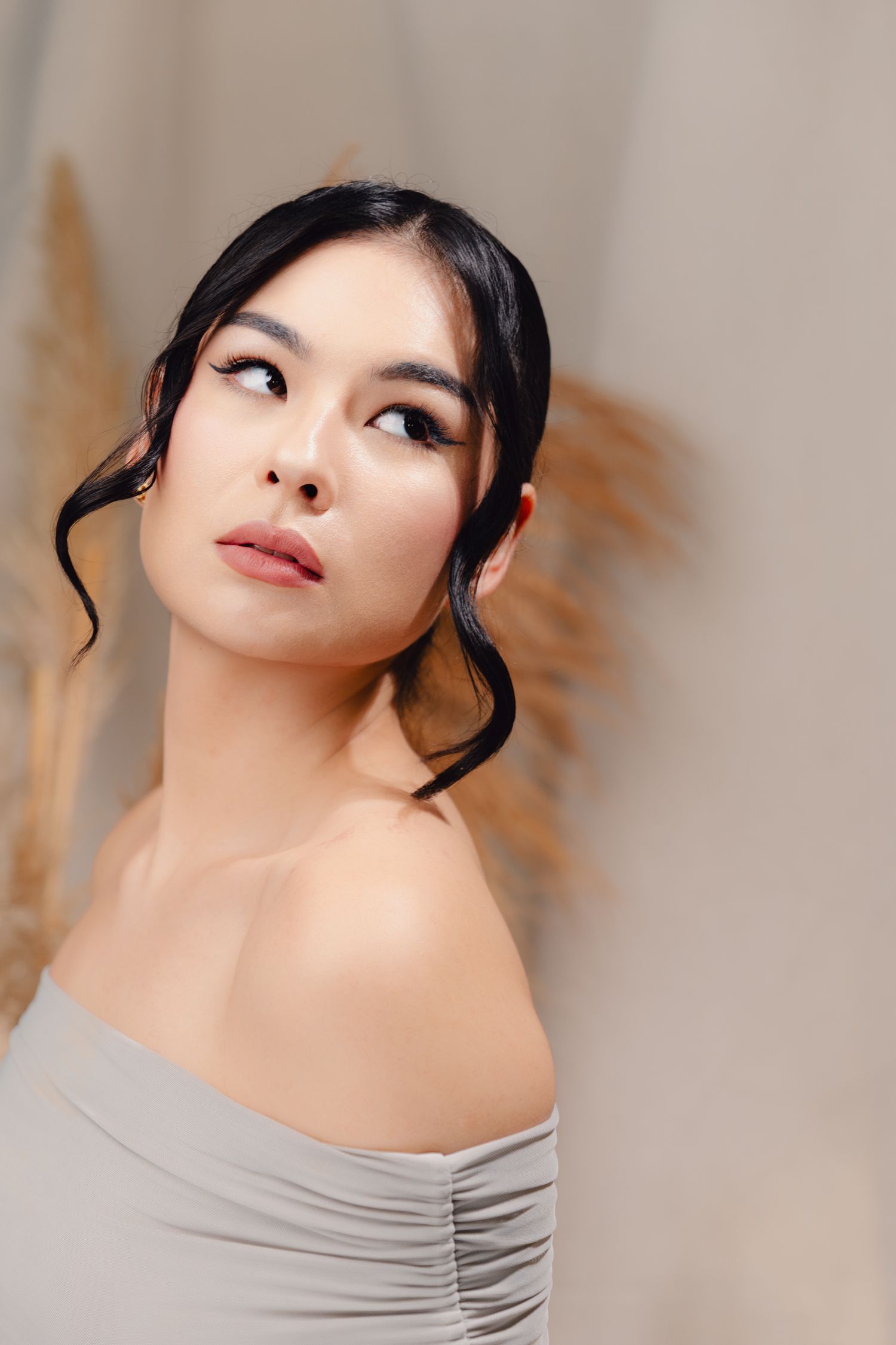 Elegant Asian woman with dark upswept hair in off-shoulder gray top, wearing natural makeup, posing against neutral background.