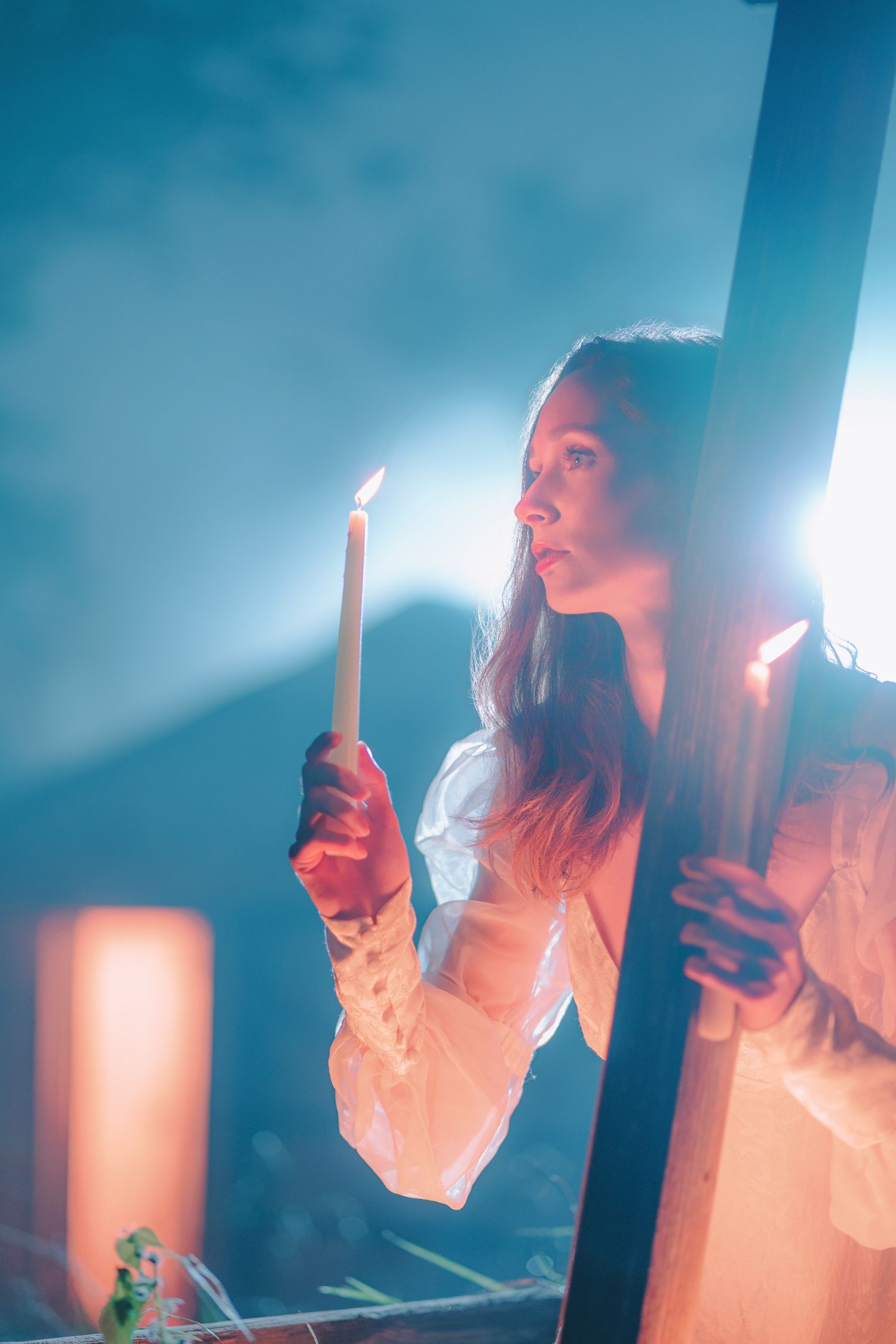 Young woman in white dress holding a lit candle with dramatic blue smoky background and warm candlelight glow.